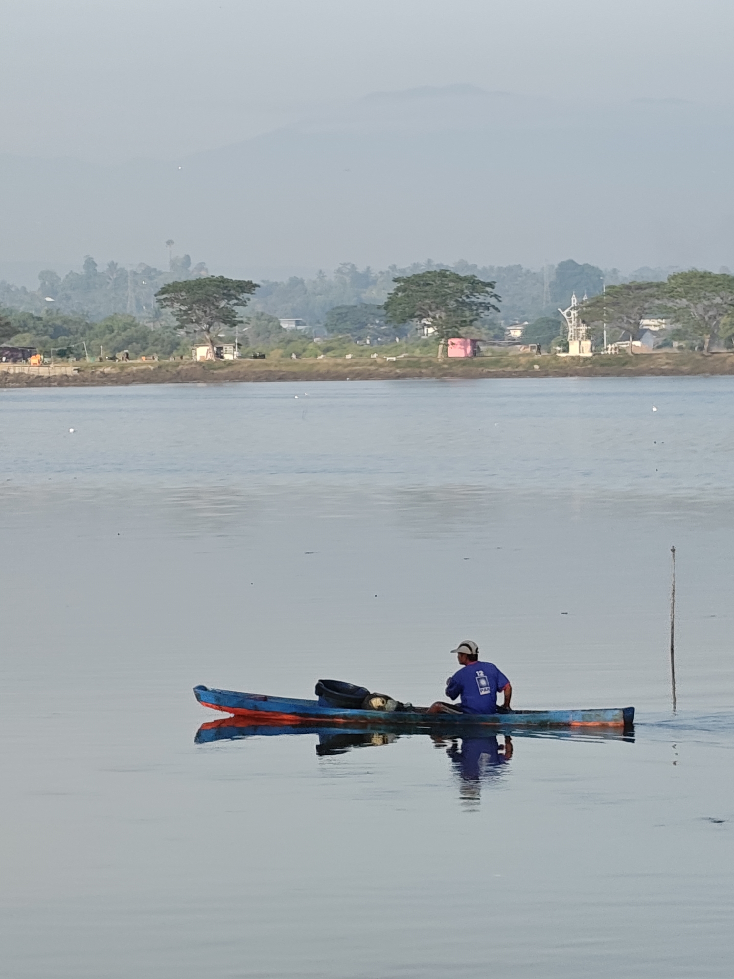 An outdoor, eye-level, medium shot captures a lone fisherman in a narrow, blue and orange boat on a calm body of water, likely the sea or a river in Lhokseumawe, Indonesia. The fisherman, wearing a blue shirt with a white logo that reads "12 PAR" and a white cap, sits facing forward, his reflection clearly visible in the still water. His hands are not visible, suggesting he may be paddling or holding something out of frame. The boat contains a dark bag behind him. A thin, vertical wooden pole stands out of the water to the right of the boat. In the background, a hazy, low-lying shoreline is dotted with trees and some structures, including what appears to be a white utility pole with wires. The sky is overcast with a light mist or fog, obscuring the distant mountains. The overall atmosphere is serene and slightly muted due to the atmospheric conditions.
