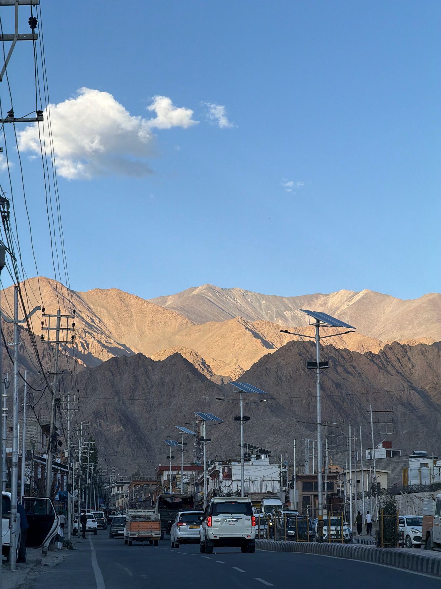 A wide, asphalt road runs through the city of Leh, India, with vehicles driving and parked on either side. The road is lined with buildings, utility poles, and streetlights, some of which are equipped with solar panels. In the background, majestic mountains dominate the skyline, their rugged slopes illuminated by the warm glow of the sun. A lone cloud drifts across the clear blue sky, adding a touch of softness to the otherwise stark landscape. The overall impression is one of a bustling city nestled within a dramatic natural setting.