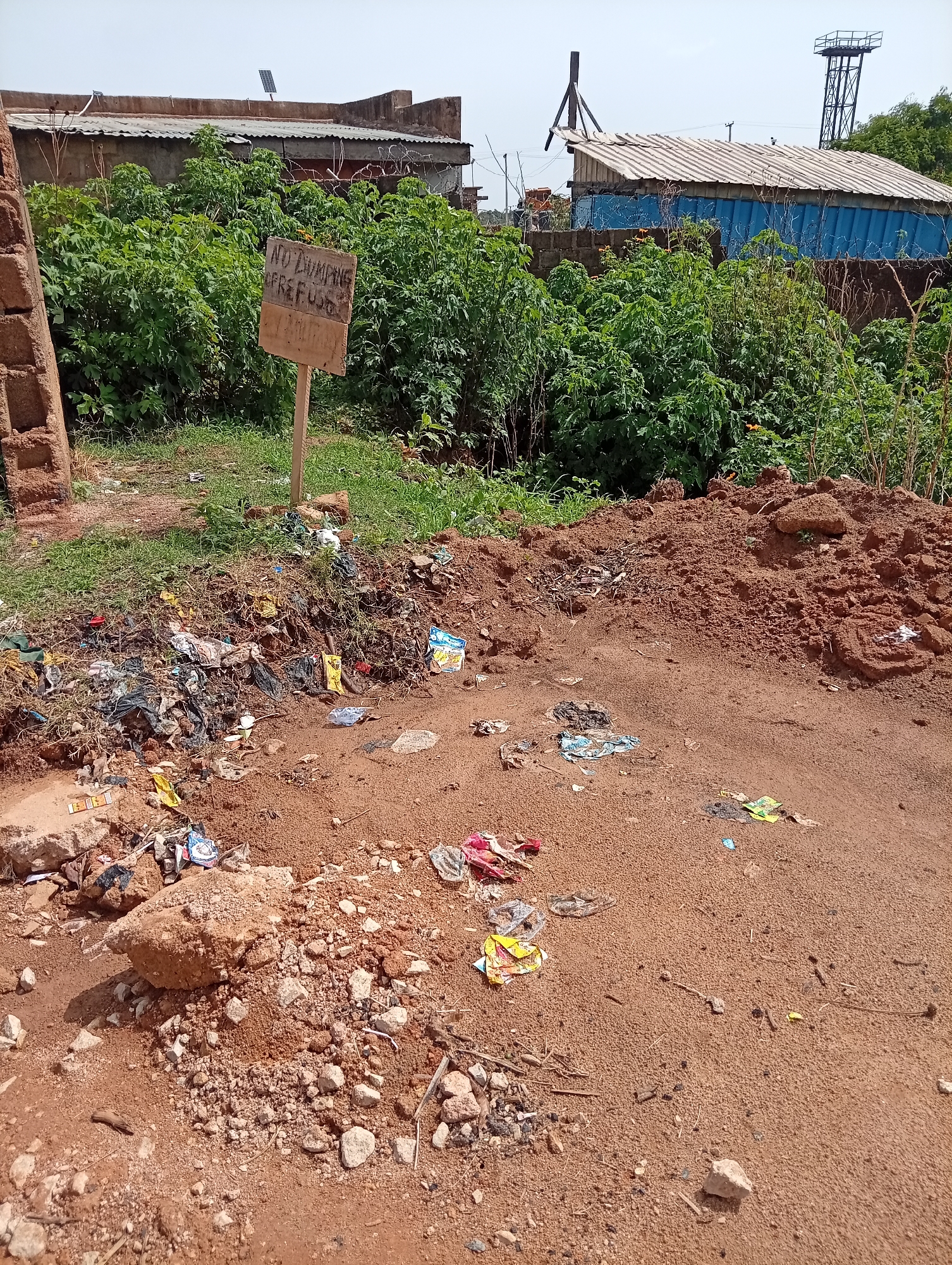The image shows a trash-strewn area in Jos, Nigeria. A handmade wooden sign reads "NO DUMPING, WREFUGE, POLLUT". Various discarded items, including plastic wrappers and debris, are scattered across the red soil. A pile of reddish-brown earth and scattered rocks lie adjacent to the trash. In the background, green vegetation partially obscures buildings with corrugated metal roofs. A metal tower is also visible. The overall scene suggests an illegal dumping site in an underdeveloped area.