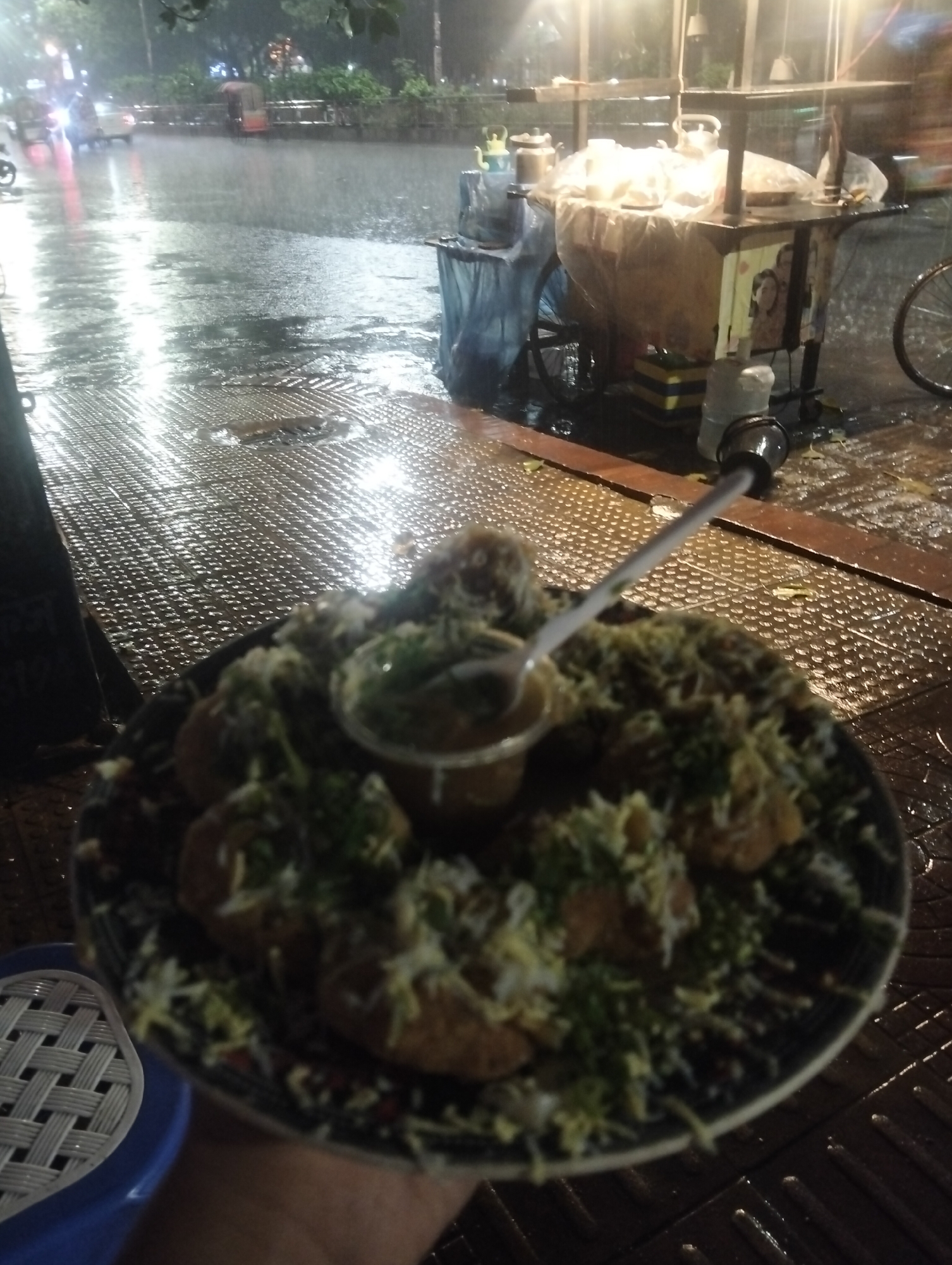 Here is a description of the image.
The image shows a plate of "pani puri" or "phuchka," a popular street food, being held in the foreground. The food is topped with shredded ingredients and a small container of sauce, along with a spoon on top. The background indicates an urban setting, likely in Dhaka, Bangladesh, given the style of the street food and overall environment. It appears to be a rainy evening. Reflections of lights are visible on the wet pavement. A street food vendor's cart is nearby.