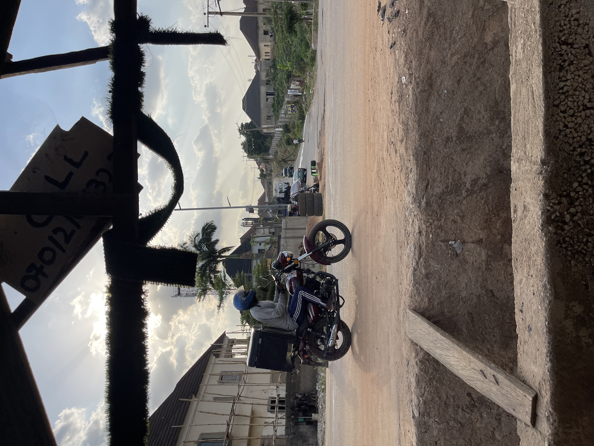 A slightly rotated outdoor shot shows a road in Abuja, Nigeria. The photograph is taken from a partially constructed building, framing the scene with scaffolding and building materials. A motorcyclist wearing a helmet is situated on the road, the vehicle being maroon with a black delivery box attached to the back. In the background, there are residential buildings, a palm tree, and other vehicles on the road. The sky is overcast with clouds. The edge of a concrete structure is visible on the right side of the frame, indicating the presence of the building's foundation or stairs.