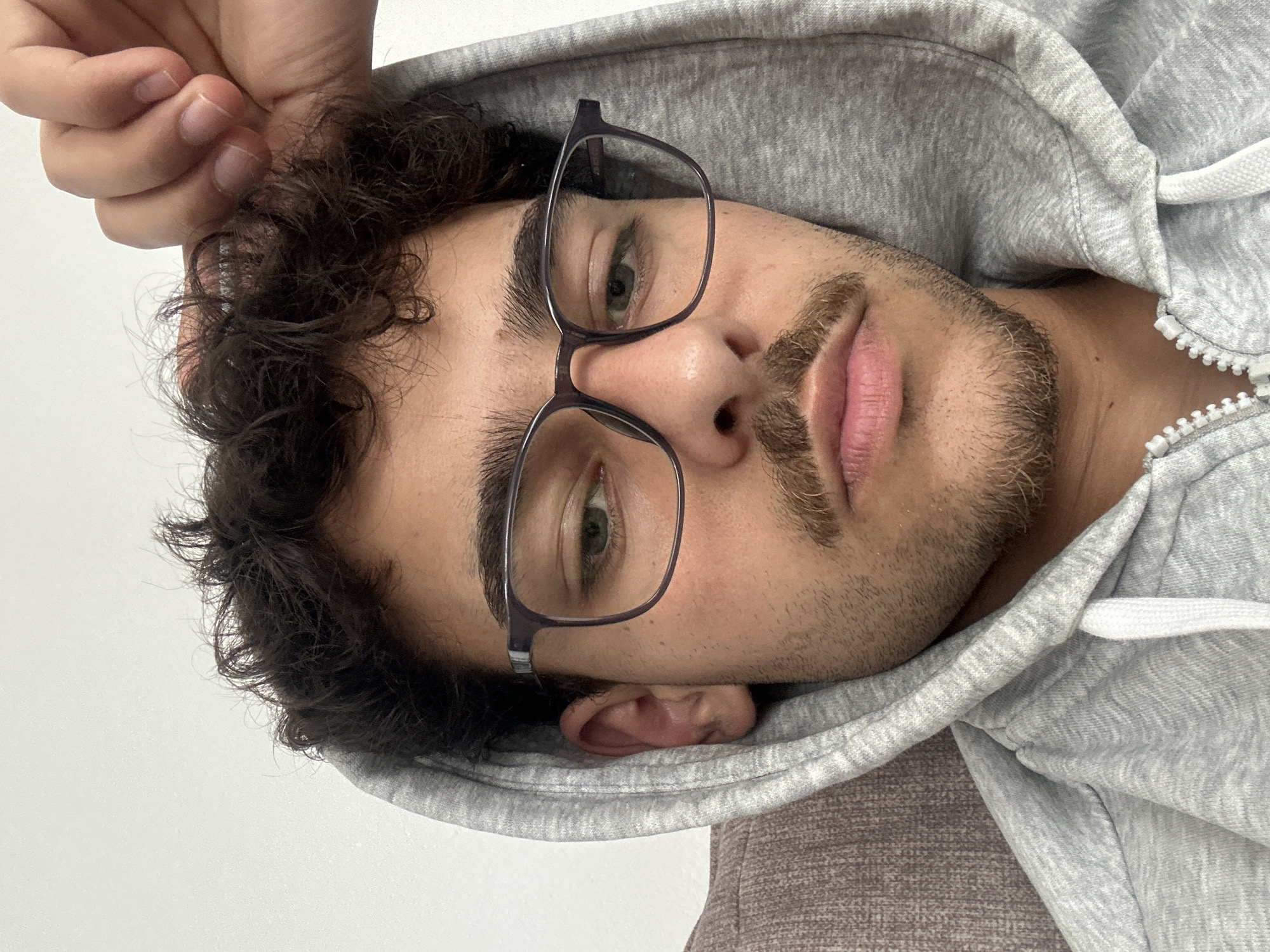 A close-up, portrait-oriented image, rotated 90 degrees counter-clockwise, features a young man with dark, curly hair, light skin, and green eyes. He is wearing dark-framed eyeglasses and has a neatly trimmed mustache and a light brown beard. The man is dressed in a heather grey hooded sweatshirt (hoodie) with the hood pulled up, framing his face. White drawstrings are visible on the hoodie.
He looks directly at the camera with a neutral or slightly pensive expression. His left hand is visible in the upper left corner of the frame, with his fingers appearing to touch or gently hold his hair. The background is predominantly plain white, suggesting an indoor setting with a simple wall or surface. A textured, possibly brown or grey, fabric surface is visible in the lower right corner, which could be part of furniture. The lighting is soft and even, illuminating his face clearly. There are no visible texts or specific location cues within the image itself, other than the provided context of Troyan, Bulgaria.