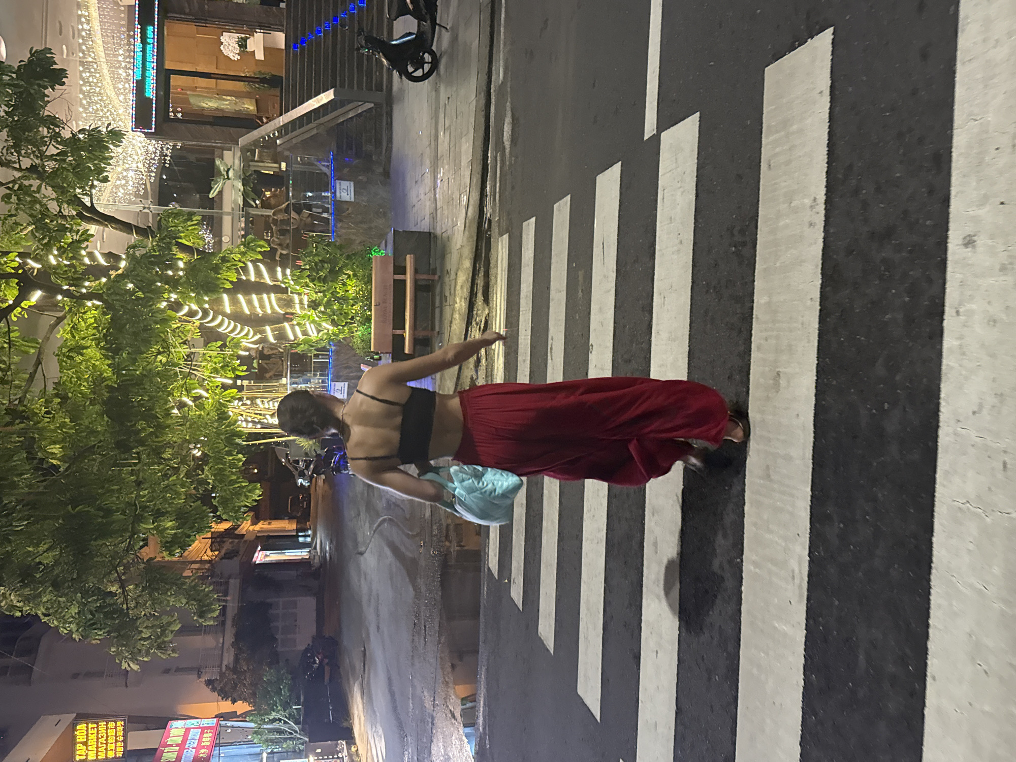The image shows a woman crossing a street at a crosswalk in Bac Nha Trang Ward, Vietnam. The woman is wearing a black top, a long red skirt, and sandals. She is also carrying a light blue bag. The street is wet and there are buildings and trees in the background. Lights are strung across the trees. A sign in Vietnamese, Russian, Chinese, and Korean is visible at the bottom left corner of the image.