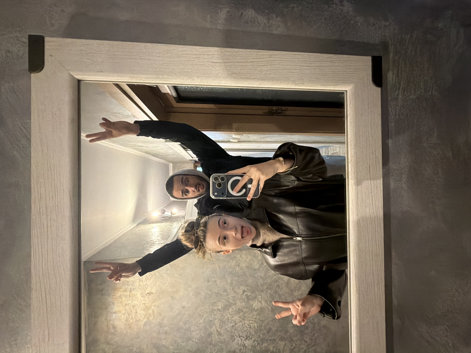 The image depicts two young adults posing for a mirror selfie in an indoor setting.
**People:**
A young adult female is in the foreground, holding a dark-colored smartphone with her right hand to capture the image. She has light-colored hair styled in a high messy bun, with sunglasses pushed up onto it. She wears a dark brown leather jacket over a light brown turtleneck sweater. Her left hand is raised, displaying a "peace" sign (V-sign). Her mouth is open with her tongue slightly extended, conveying a playful expression. A ring is visible on her right ring finger and a bracelet on her right wrist.
Behind her, a young adult male is visible, with a dark hoodie pulled over his head. He has facial hair and also has his mouth open with his tongue slightly extended, mirroring the female's playful expression. Both of his hands are raised above his head, each forming a "peace" sign (V-sign).
**Objects:**
The subjects are reflected in a rectangular mirror with a wide, light-colored wooden frame. The frame features dark, possibly metallic, corner accents on the upper right and lower right visible portions. The female is holding a dark-colored smartphone with multiple camera lenses visible on its back.
**Environment:**
The background reflected in the mirror appears to be a hallway or corridor. The walls have a textured, possibly stucco or metallic-effect, grey finish. A doorway with a dark panel is visible on the right side of the reflection. A light fixture with multiple bulbs is present on the upper right of the reflected background. The wall surrounding the mirror exhibits the same textured grey finish as the reflected background walls.
**Location in Plovdiv, Bulgaria:**
The specific location within the city of Plovdiv, Bulgaria, cannot be determined from the visual evidence provided; it is an interior space, likely a private residence or commercial establishment.