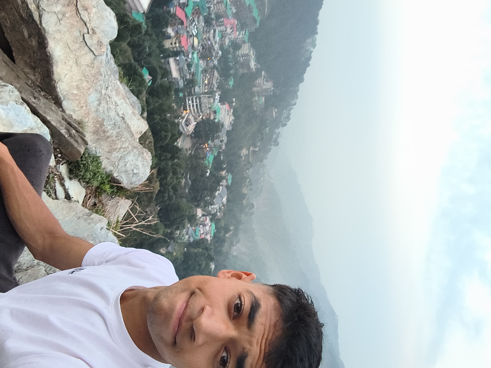 The media file shows a person taking a selfie while sitting on a rocky ledge overlooking a town nestled in a valley. The person, a young male with dark hair and eyes, is wearing a white t-shirt and dark pants. He is smiling and looking directly at the camera.
The town below is densely populated with buildings of various colors and sizes, including many with red and green roofs. The town is surrounded by lush green trees and mountains. The sky is overcast and hazy, suggesting either early morning or late afternoon, or possibly misty weather conditions. The overall impression is a scenic and perhaps remote location.
The setting appears to be mountainous terrain, with the rocky ledge in the foreground providing a sense of height and perspective. The presence of trees and the general landscape suggest a natural environment. The activity captured is a personal moment of taking a photograph, likely to capture the view and the experience of being in this location. There is no visible text in the image. The location context of Bhagsu, India, further supports the interpretation of a mountainous, possibly religious or tourist destination.