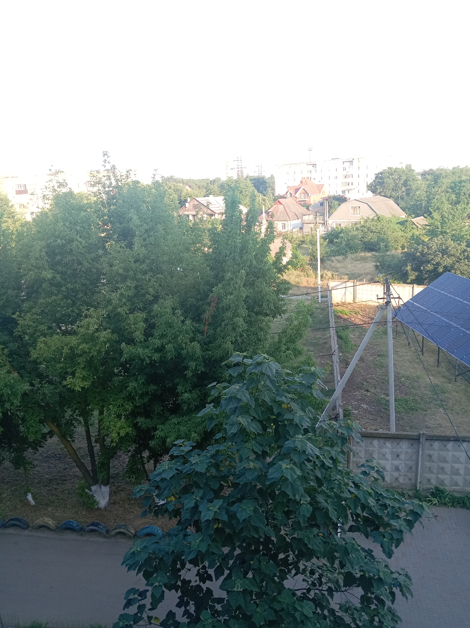 The image shows a view of a residential area in Velykodolynske, Ukraine, likely during daylight hours. In the foreground, a large tree with dark green leaves partially obscures the view. To the right, a solar panel array is visible, angled towards the sky. Beyond the solar panels, a concrete fence runs horizontally across the midground, separating the foreground from a grassy area. In the background, several houses with pitched roofs and one larger, multi-story building are nestled amongst lush green trees. The sky is bright, almost blown out, suggesting a sunny day. There are no people or animals visible in the scene.