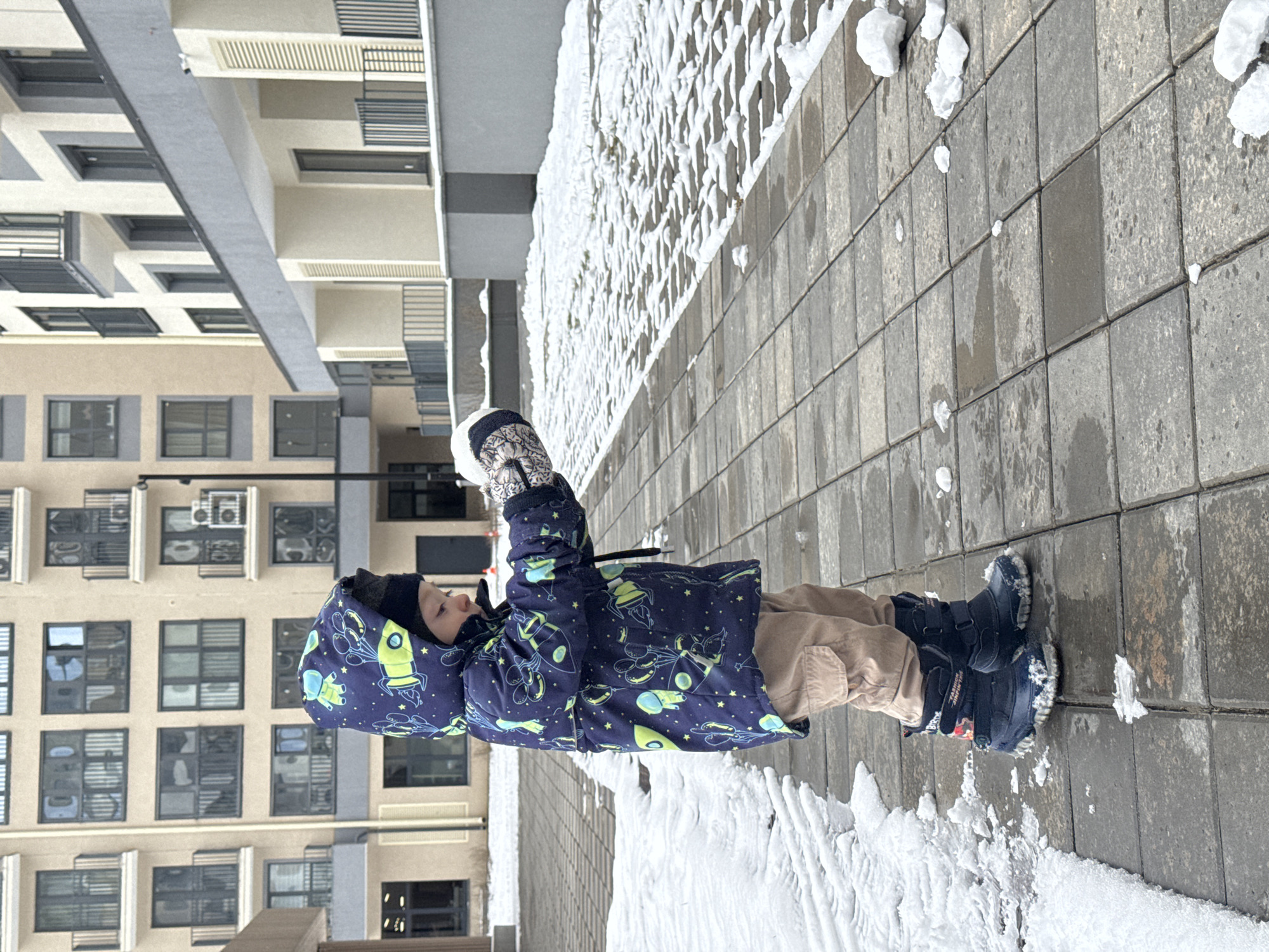 A child in winter attire stands on a gray, paved sidewalk with patches of snow. The child wears a navy blue jacket with a rocket and balloon pattern, beige pants, and dark blue boots. They also have patterned mittens and a black beanie. The child appears to be holding a ball of snow. Behind the child is a modern building with multiple windows and balconies, likely an apartment complex. A wall is covered in snow. The city is Izhevsk, Russia.