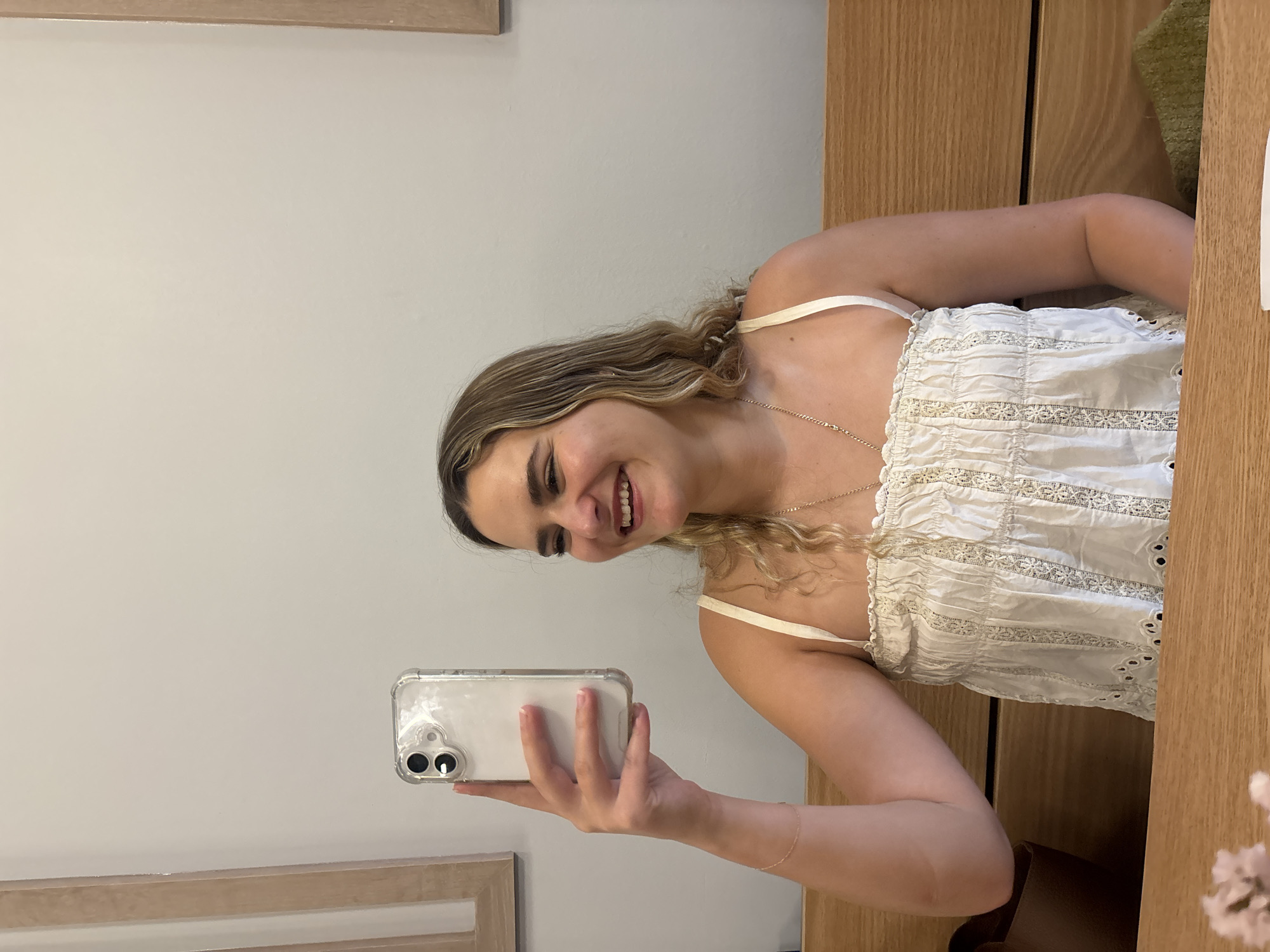 A fair-skinned young woman smiles while taking a selfie with an iPhone. She is wearing a white, sleeveless top with eyelet trim. She has blonde hair and is wearing a gold necklace. The background consists of a light gray wall and a wooden cabinet. The location appears to be indoors. The image does not contain indicators relating it to Puebla, Mexico.