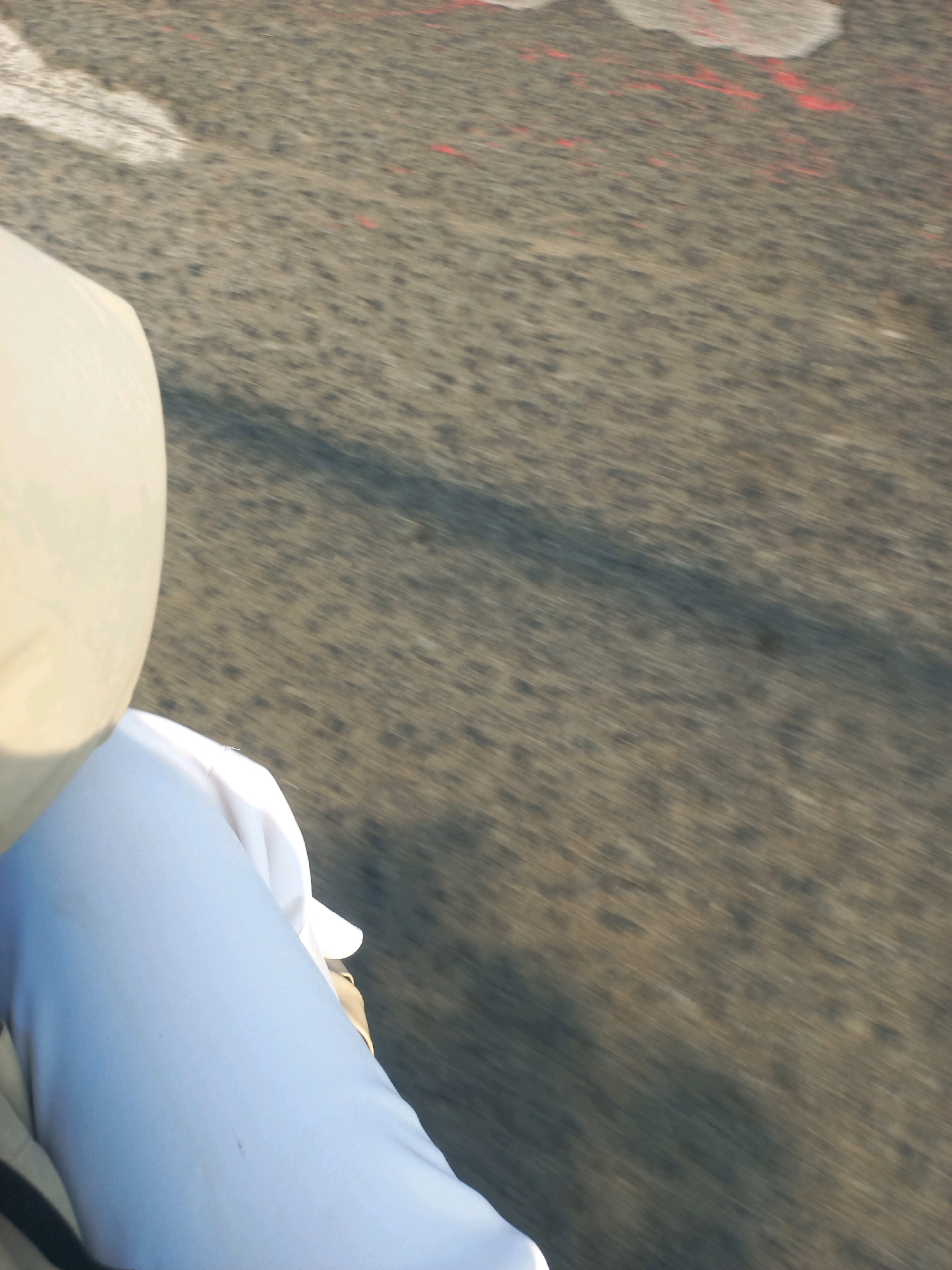 A first-person perspective shot captures a person in white clothing, possibly a thawb, sitting on a road in Baturetno, Indonesia. The person's left side is visible, with a beige garment draped over their shoulder. The road, likely asphalt, is textured and shows signs of wear, with faint red markings and a white line visible on the left side. A shadow stretches across the road, indicating sunlight. The image is blurry, suggesting movement, likely from a vehicle.