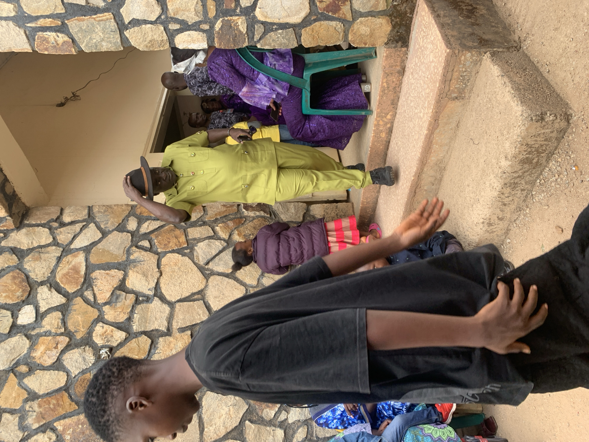 On the steps and in front of a stone building in Jos, Nigeria, a group of people are gathered. An individual in a green suit and a hat stands near the doorway, with several people seated or lying on the steps behind him. A woman in purple clothing sits on a green plastic chair. In the foreground, a person in a black shirt appears to be gesturing, with other people sitting on the ground near their feet. The environment appears to be a public space with stone architecture.