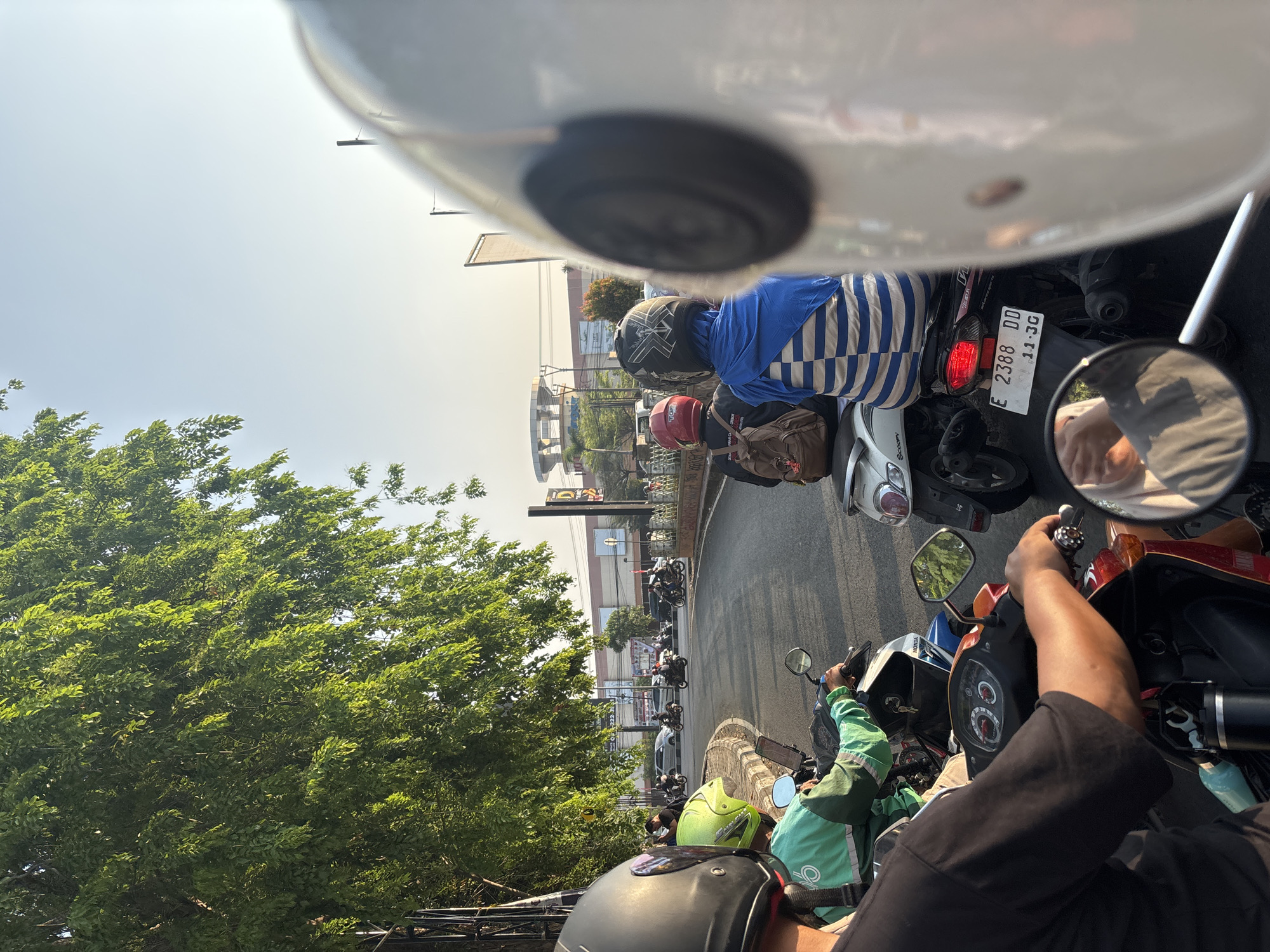 In a street scene in Cirebon, Indonesia, a group of motorcyclists are stopped at an intersection, likely due to traffic. The foreground is dominated by the back of a white motorcycle helmet, partially obscuring the view of the rider and their handlebars. To the left, a person wearing a green jacket with the "GoFood" logo rides a motorcycle, their neon green helmet visible. Behind them, a motorcycle with a blue and white striped passenger sits at the rear. The road curves to the right, lined with trees on the left, casting dappled sunlight on the scene. In the background, a large building with a distinctive circular facade, possibly a shopping mall, stands under a clear sky. The overall impression is of a typical urban commute in Indonesia, with motorbikes being the primary mode of transportation.