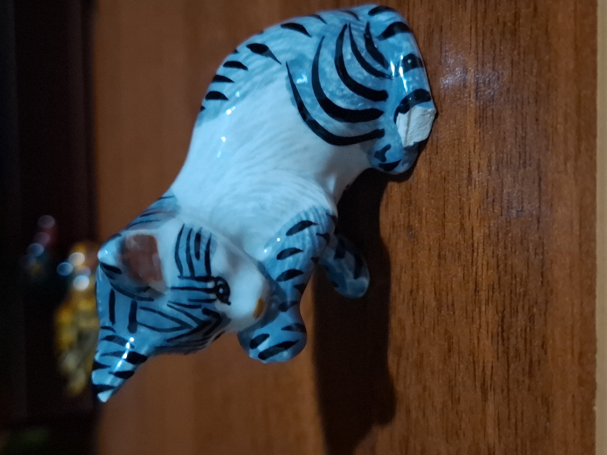 A small, ceramic figurine of a light blue tabby cat with black stripes sits on a wooden surface. The cat is in a crouched position with its front paws raised to its face. The background is dark and out of focus, with some blurred lights visible. The scene is indoors, on a wooden table. There are no people present. The image does not provide any information about the location in Almaty, Kazakhstan.