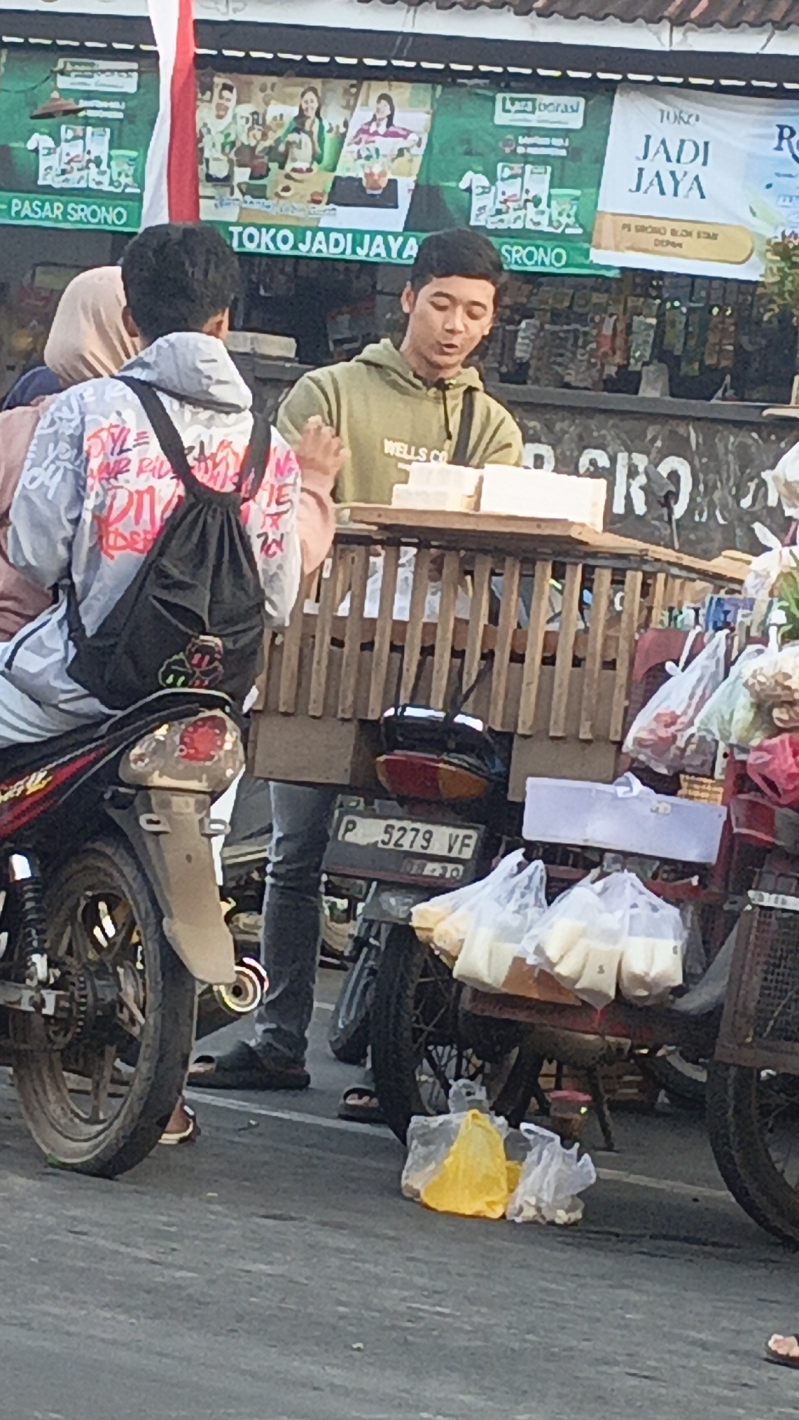 In Srono, Indonesia, a young man wearing a green hoodie with "WELLS CO" on it stands behind a wooden cart laden with food items. He appears to be speaking to someone off-camera. In front of him, on the cart, are several white rectangular packages, possibly food. To the left, a person wearing a jacket with colorful graffiti and a black backpack is seated on a red motorcycle. Behind the young man, a banner displays text including "PASAR SRONO," "TOKO JADI JAYA," and "SRONO." The cart is overflowing with plastic bags containing various goods. The scene appears to be a market or street stall setting with motorcycles parked nearby and people engaged in commerce.