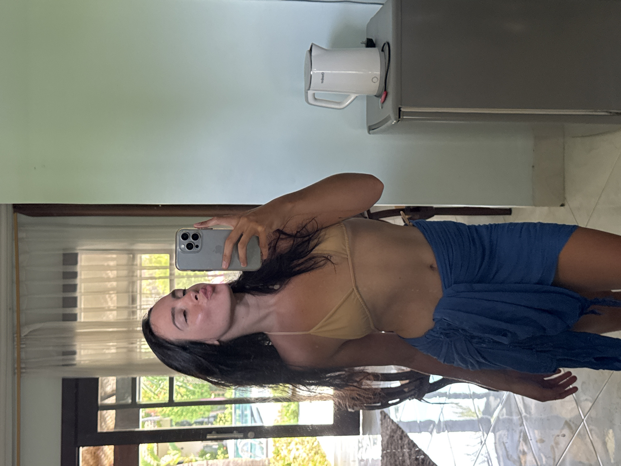 A woman is taking a mirror selfie in a room. She is wearing a tan bikini top and a blue sarong. Her hair is dark and long, and she is making a kissing face. In the background, through a doorway, a swimming pool and palm trees are visible. To her right, a white electric kettle sits on top of a gray mini-fridge. The room has tiled floors and light-colored walls.