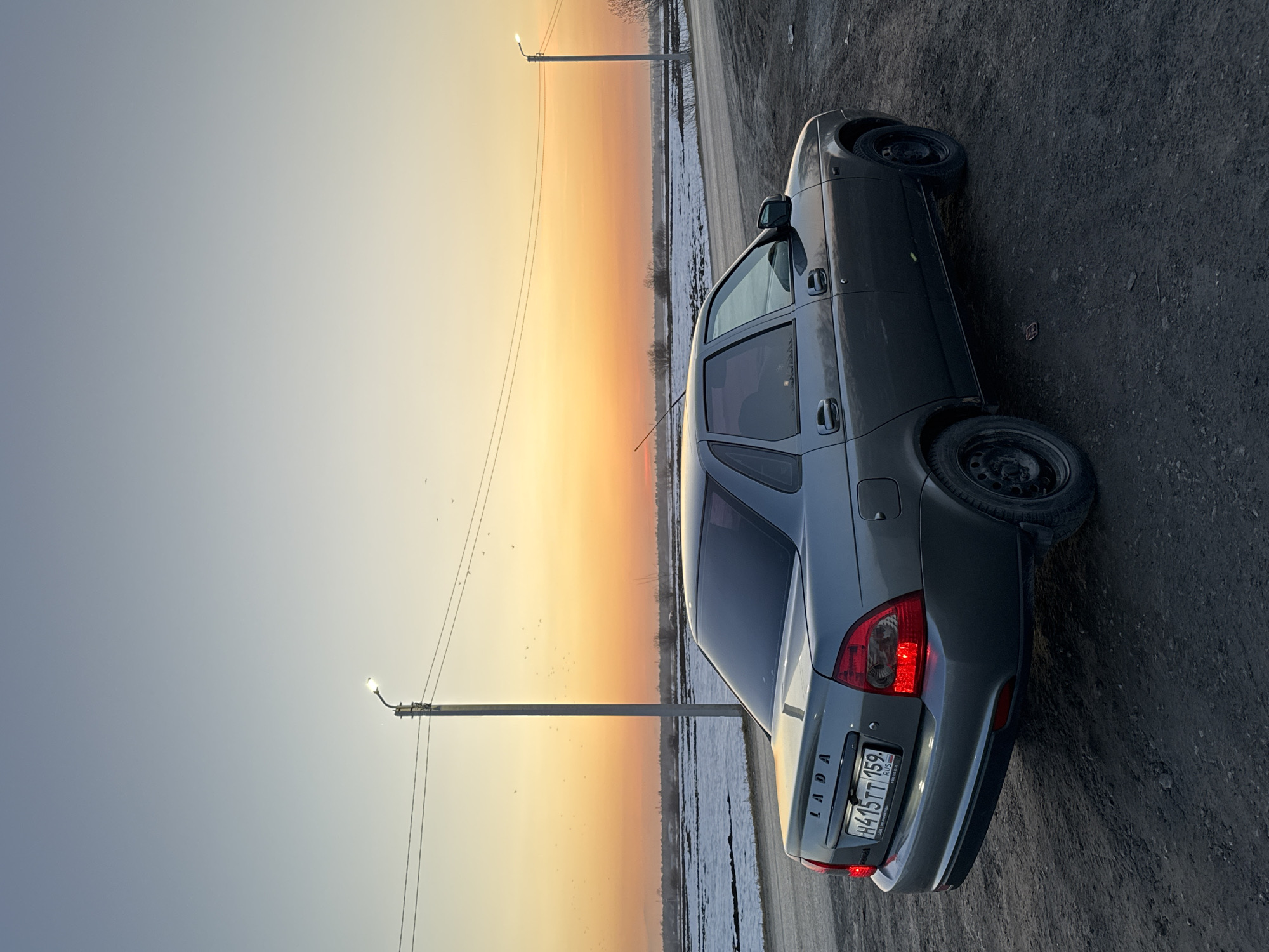 A medium shot depicts a gray Lada sedan parked on the shoulder of a road, presumably in Кондратово, Russia, based on the Russian license plate and the "Lada" badge on the car's rear. The car is positioned at a slight angle to the camera. The background features a twilight sky with a gradient of colors transitioning from a pale blue to a warm orange. Two utility poles are visible, extending upwards into the sky. The ground adjacent to the road appears to be a mix of dirt and some vegetation, potentially with remnants of snow or frost. The overall setting seems to be a rural or semi-rural roadside scene at dusk or dawn.