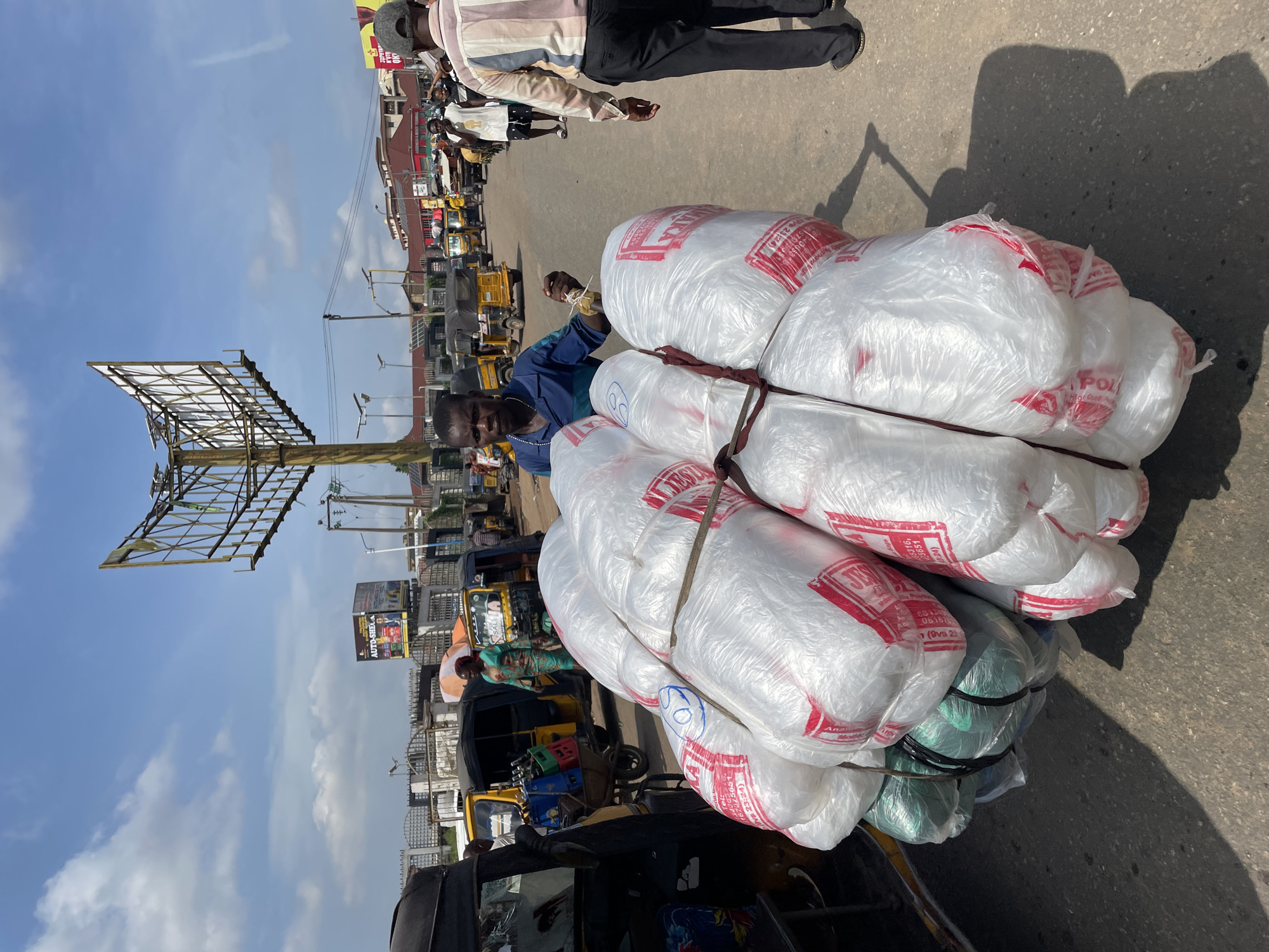 This media file depicts a bustling street scene in Onitsha, Nigeria. The primary subjects are people and objects, with a focus on a man in a blue shirt tying up large, white plastic bags filled with goods. These bags are secured with ropes and appear to be stacked high, possibly on a cart or vehicle. The man is looking towards the camera, appearing engaged in his task.
The setting is an outdoor street, likely a market area, given the presence of other vehicles and people. Yellow auto-rickshaws, a common mode of transportation in Nigeria, are visible in the background, along with other people going about their day. A large, partially obscured billboard stands prominently on the left side of the frame, advertising "AUTO-SHEA". The weather appears to be clear and sunny, with visible shadows on the asphalt road, suggesting it is daytime.
The overall activity is the transportation of goods in a busy urban environment. The image captures a moment of everyday commerce and movement in Onitsha. Text visible on the white bags includes "JES," "POL," and what appears to be contact or product information. The scene conveys a sense of active street life and economic activity.