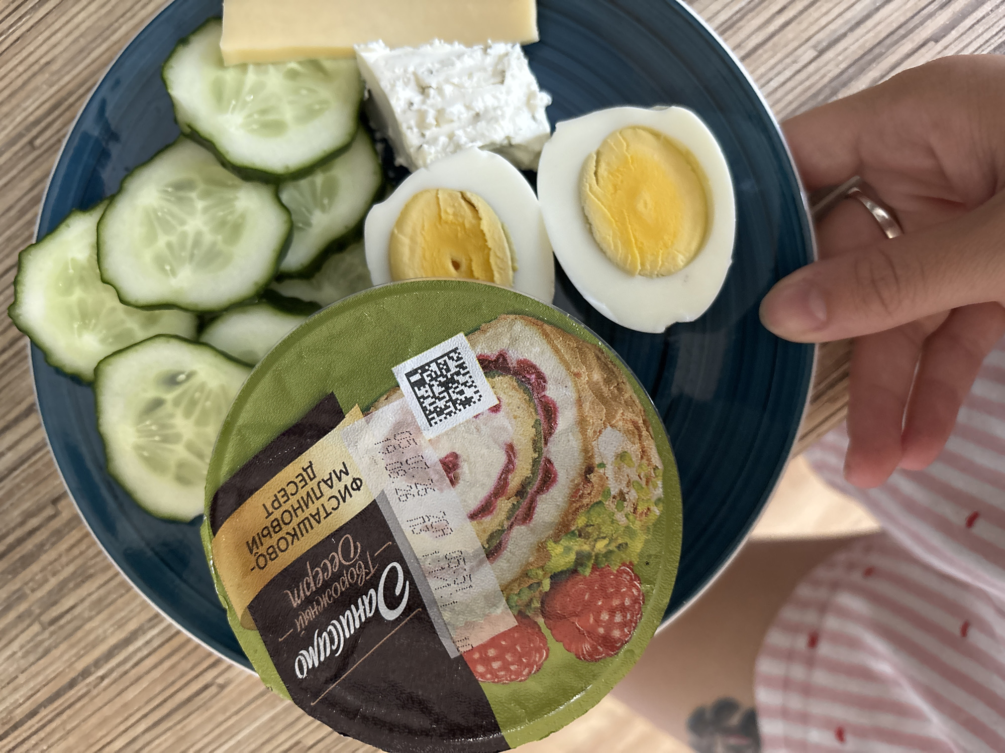 This is a close-up, overhead shot of a dark blue plate filled with breakfast items. The plate contains sliced cucumbers, two hard-boiled eggs, a wedge of yellow cheese, and a block of white cheese. A round, green-lidded container with a label is placed on top of the other food items. The label features a picture of raspberries and what appears to be a dessert. It also has Russian text, including "САМСИНО" and "Творожный десерт". Beneath the label, a crumpled receipt is partially visible, displaying dates and numbers. The background shows a light-colored wooden surface. A person's hand with a silver ring is visible at the bottom right, holding the plate. Their legs, clad in pink and white striped shorts with small red accents, are also in the frame at the bottom left. The scene is likely in a residential setting. No specific location within the city of Naberezhnye Chelny, Russia, is identifiable from the image.