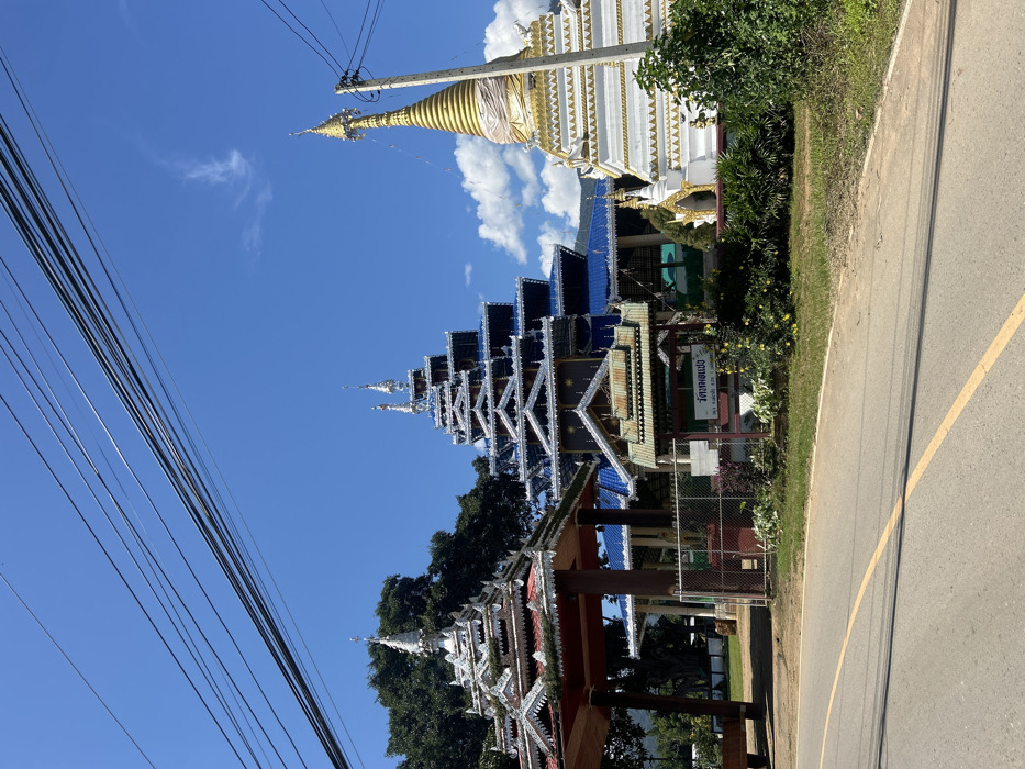 An exterior, static daytime image depicts a Buddhist temple complex in Ban Mai Saha Sampan, Thailand, under a clear blue sky with minimal white clouds. The scene is viewed from across a paved road.
The foreground features a two-lane asphalt road with a single solid yellow centerline. To the right of the road, a dirt shoulder transitions into a strip of green grass and low-lying shrubs. A low, brown chain-link fence partially delineates the temple grounds from the roadside. Behind the fence, a small wooden sign with a brown tiered roof displays text in Thai script, flanked by yellow flowering plants.
The midground and background are dominated by three distinct temple structures:
1. **Left structure:** A multi-tiered pavilion or entrance gate with intricate white and silver decorative elements and red trim on its roofs, topped with a spire. A large green tree is visible directly behind it.
2. **Central structure:** A prominent, multi-story main temple building featuring numerous tiered blue roofs and extensive white and silver ornamentation. Multiple spires crown its peak.
3. **Right structure:** A white chedi (stupa) with multiple tiered levels, encircled by gold banding and topped with a conical gold spire. It stands next to a grey utility pole.
Distant green hills are visible on the horizon behind the temple structures. Multiple black electrical or communication wires stretch diagonally across the upper portion of the image, obscuring parts of the sky and the temple spires. No individuals are present in the image.