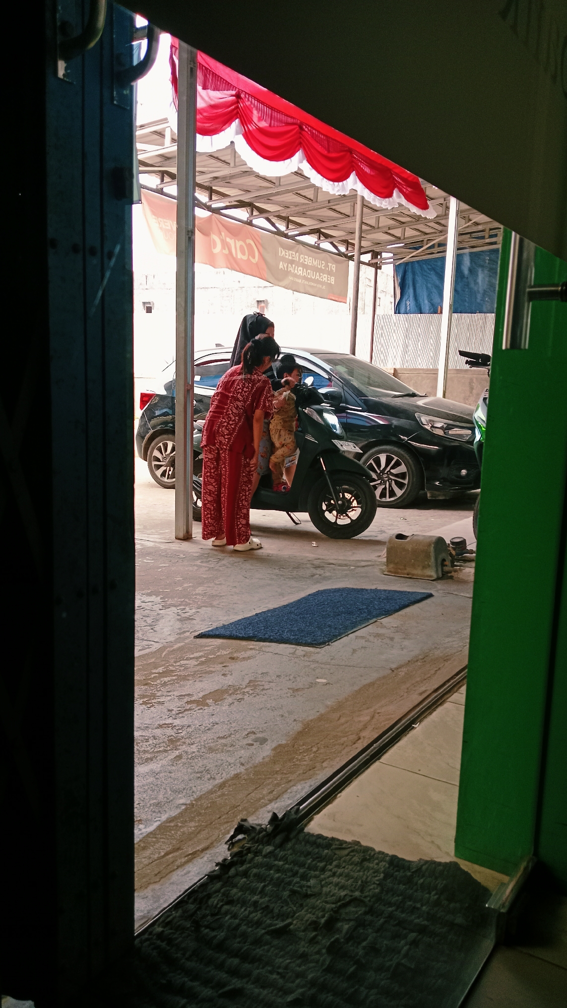 In Handil Bakti, Indonesia, an adult woman wearing a red and white patterned garment and a headscarf is standing next to a black scooter. She is leaning towards the scooter, appearing to interact with a child who is seated on the scooter's passenger seat. Another adult, also wearing a headscarf, is standing behind the first woman, partially obscured. A black car is parked behind the scooter. The scene takes place outdoors under a covered structure adorned with red and white fabric. The ground is concrete and appears damp in some areas. A blue mat is placed on the ground in front of the scooter. The overall setting suggests a workshop or a service area.