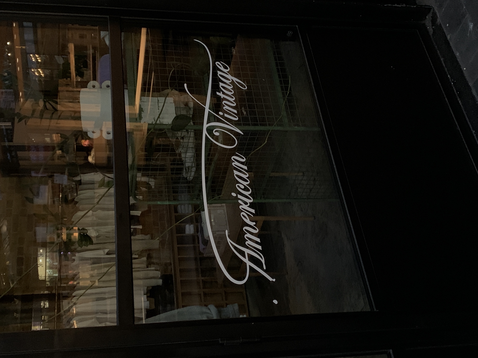 This media file depicts a shop window at night in London, United Kingdom. The dominant feature is the store's name, "American Vintage," rendered in elegant white script and oriented vertically on the glass pane.
Through the reflective glass, the interior of the shop is dimly visible. It reveals wooden shelving units, racks of folded or hung garments, and some green plants. A unique white object, possibly a ceramic or plastic decor item with circular 'eyes,' is prominently displayed inside. The interior lighting casts a warm, soft glow, contrasting with the dark exterior.
The right side of the image shows a dark, solid structure, likely a door or an adjacent wall of the building. The window's surface also reflects blurred elements of the urban environment outside, including distant lights from other buildings or street fixtures. The overall scene is static, with no people or active events taking place, suggesting a quiet, late-evening or nighttime setting. The window is framed by dark borders, emphasizing its role as a storefront display.