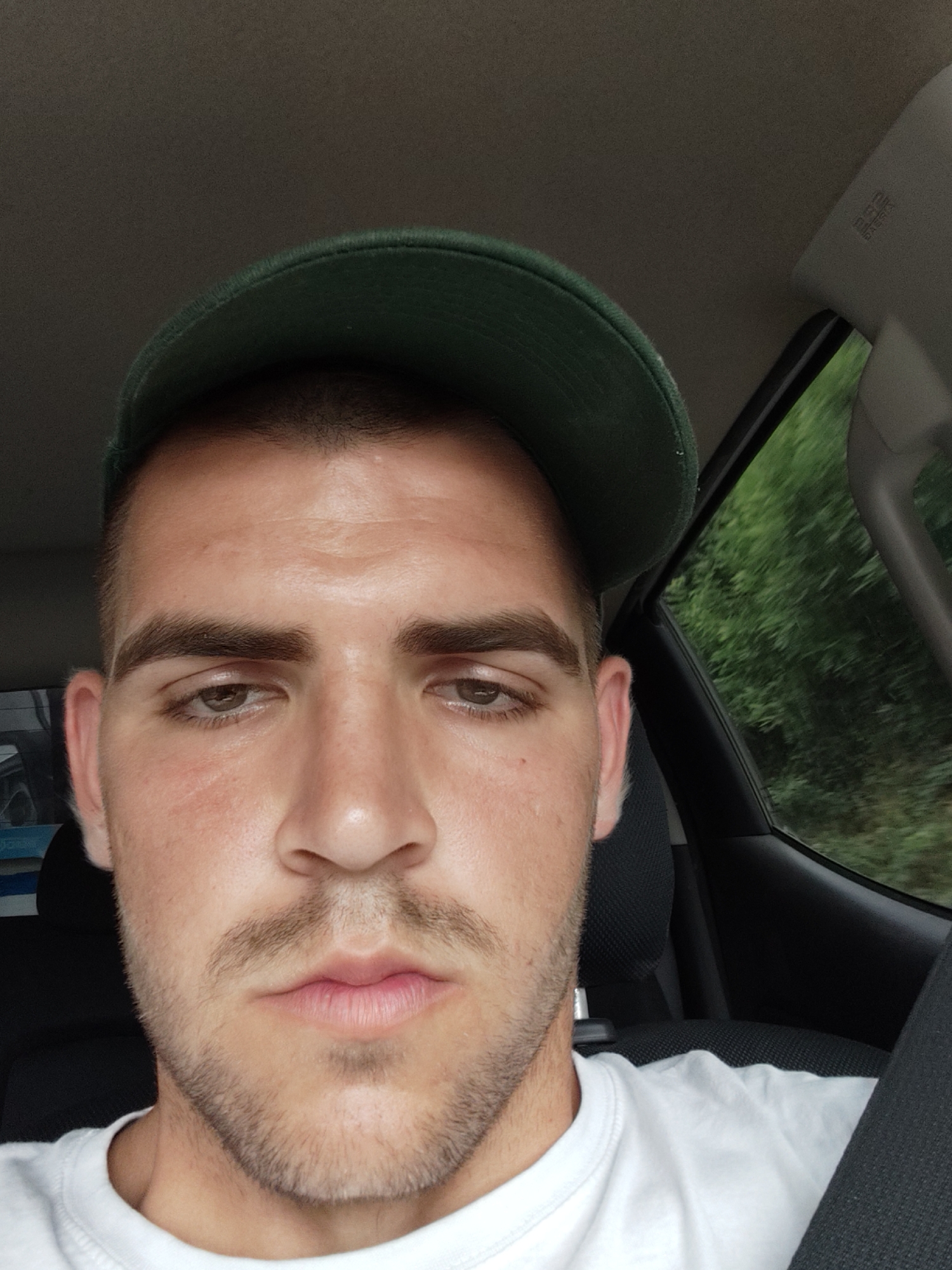 A close-up, front-facing shot shows a young Caucasian male wearing a green baseball cap tilted forward and a white t-shirt. He is in the driver's seat of a car, with the interior visible behind him. The background shows blurred green foliage, suggesting the car is in motion. The man has short brown hair, dark eyebrows, and light brown eyes. He has a light beard and a neutral expression. The lighting is natural, coming from the side and front. The image is in focus and sharp, capturing the details of his face and clothing.
