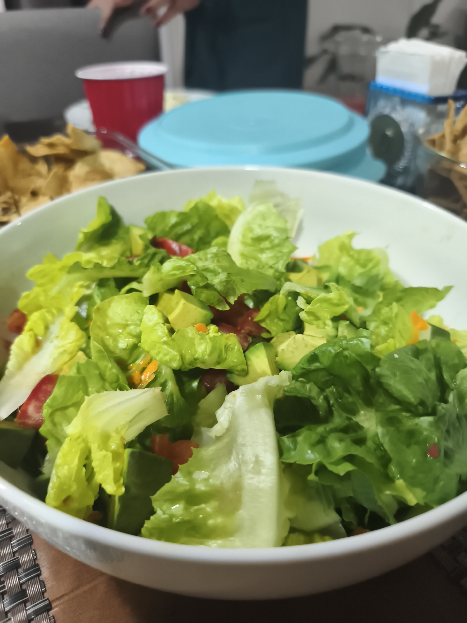 The image shows a white bowl filled with a salad. The salad appears to contain lettuce, diced avocado, and red bell pepper. The bowl sits on a brown surface with a woven pattern. In the background, there's a red plastic cup, a light blue lid, a container of tissues, and a bowl of tortilla chips. People can be seen in the background out of focus. The location is unknown, and there is no indication that the image was taken in Mexico City, Mexico.