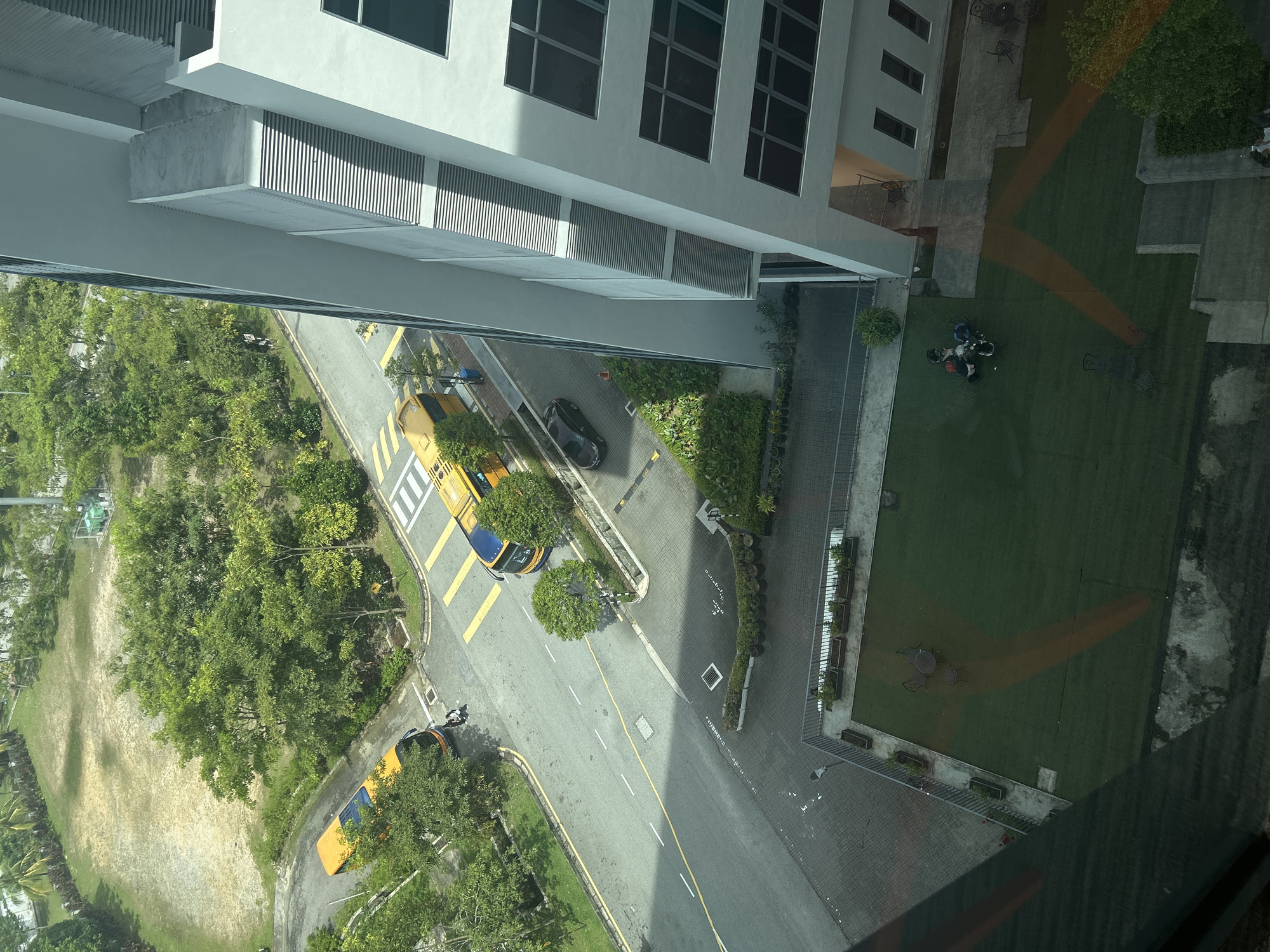 The image shows an aerial view of a street scene in Kuala Lumpur, Malaysia. A multi-lane road with yellow and white lane markings is visible, with yellow school buses parked along the side. A black car is parked on the side. Adjacent to the road, there is a grassy area with trees and shrubs. Part of a building with multiple windows is visible at the top. In the top right, there is a lawn. A table and four chairs sit on a patio area. Three people in a group are standing together. The photo is taken from inside a building, looking out and down, with some reflections visible in the glass.