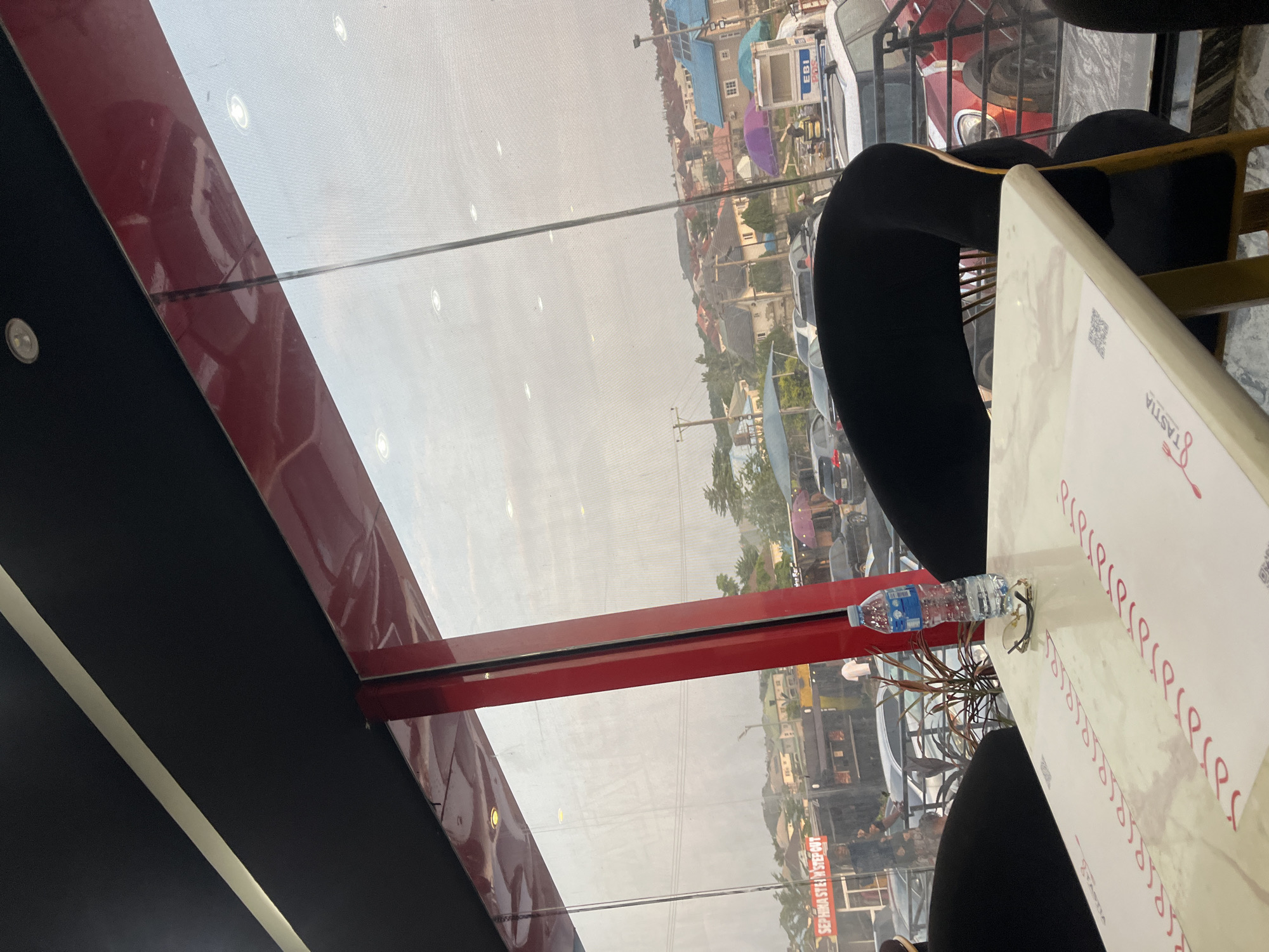 The image presents an interior perspective from a commercial establishment, likely a cafe or restaurant, overlooking an urban landscape in Galadimawa Village, Nigeria. In the immediate foreground, a light-colored, possibly marble-patterned table is visible, upon which rests a plastic water bottle with a blue label and a pair of sunglasses. A paper placemat on the table features red, stylized script and a partial "TASTIA" logo with a fork icon, along with a QR code. Portions of two black, upholstered chairs with wooden or metallic frames are seen around the table. The interior ceiling is black, incorporating a linear light fixture and several recessed circular spotlights, some of which cast reflections on the window.
A large, red-framed glass window dominates the view, revealing the exterior. Through the window, a street scene unfolds with parked and moving vehicles. On the left, a building displays signage reading "SEPHINA STEIN STEP OUT." Further right, another structure features "EBI POS" prominently. Beyond the immediate street level, a dense cluster of residential buildings with varied roof colors, including blue and reddish-brown, extends up a gentle slope. Interspersed among the buildings and along the street are various trees and sparse vegetation. The sky appears overcast or hazy, providing diffused natural light to the outdoor scene. No specific human actions or interactions are discernible beyond the general presence of people and vehicles on the street.
