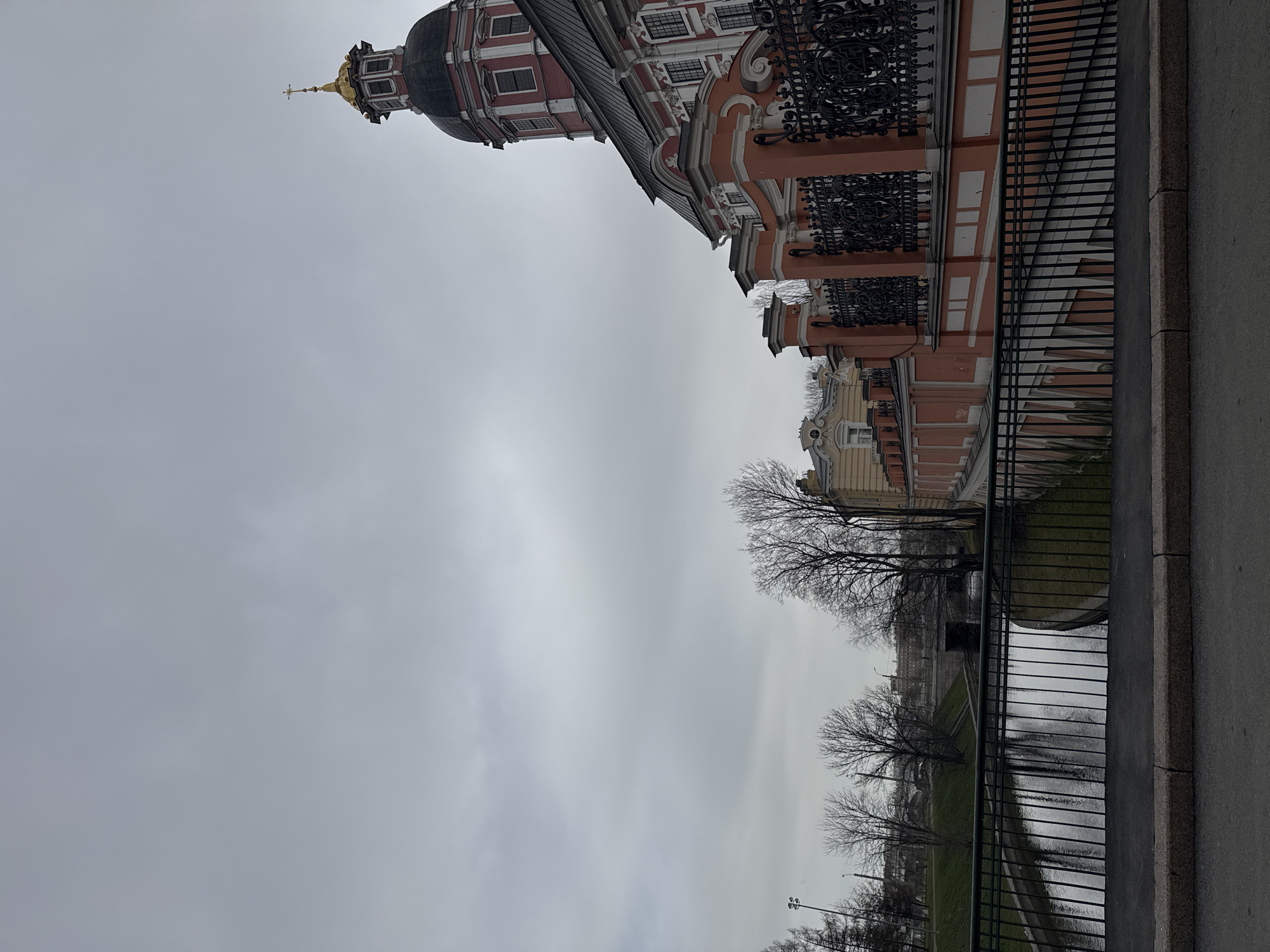 The image shows a view in Saint Petersburg, Russia, looking up at a building with a distinctive architectural style. The building has a red and white facade, intricate wrought-iron balconies, and a golden dome topped with a cross. A narrow river or canal is visible with a black railing bordering it. Bare trees line the banks, and across the river is a grassy area. The sky is overcast, indicating a cloudy day. A grey sidewalk is in the right foreground. The image has been rotated, making the composition appear upside down.