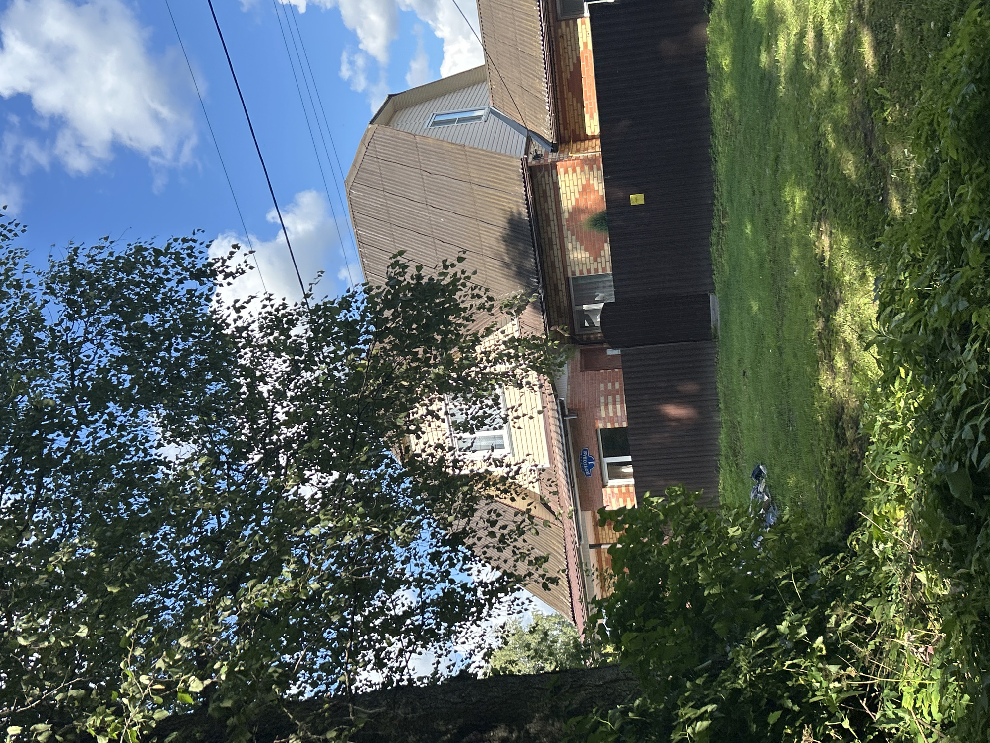 In Kalitsino, Russia, on a sunny day with scattered clouds, a two-story brick and siding house is visible. The house has a corrugated metal roof and several windows. A dark brown corrugated metal fence runs along the front of the property, separating it from a grassy yard. A mature tree with dense green foliage partially obscures the left side of the house. In the foreground, lush green bushes and vegetation are present. Near the fence and on the grass, there is a pile of dark objects, possibly trash or discarded items. A small sign with blue lettering is affixed to the front of the house above an entrance. Power lines are visible against the blue sky.