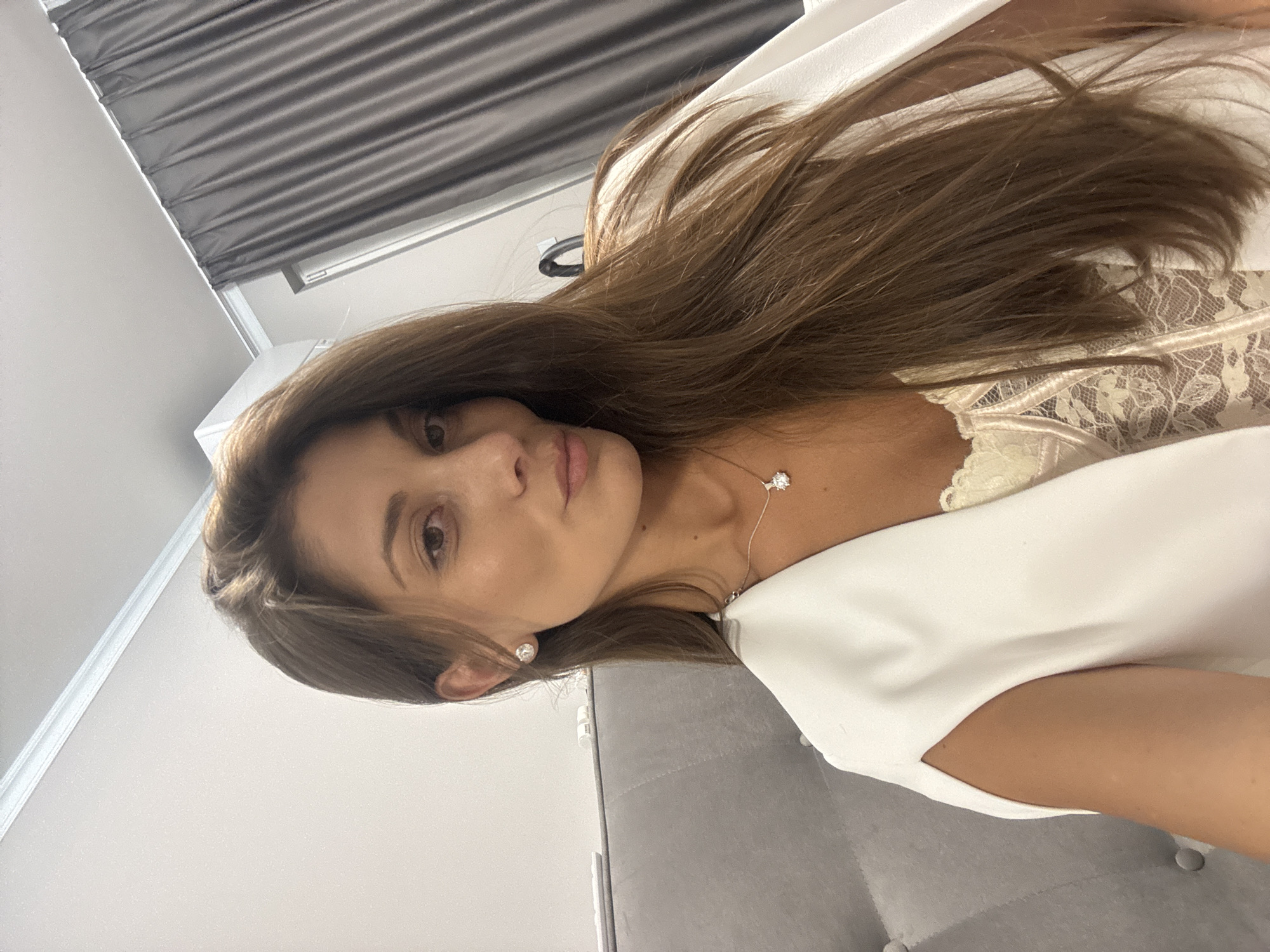 A woman with light brown hair and a fair complexion is pictured indoors. She is wearing a white dress with a lace top and a diamond necklace and earrings. The background includes a grey curtain and a light grey wall. The location in the city of Burgas, Bulgaria is not discernible from the image.