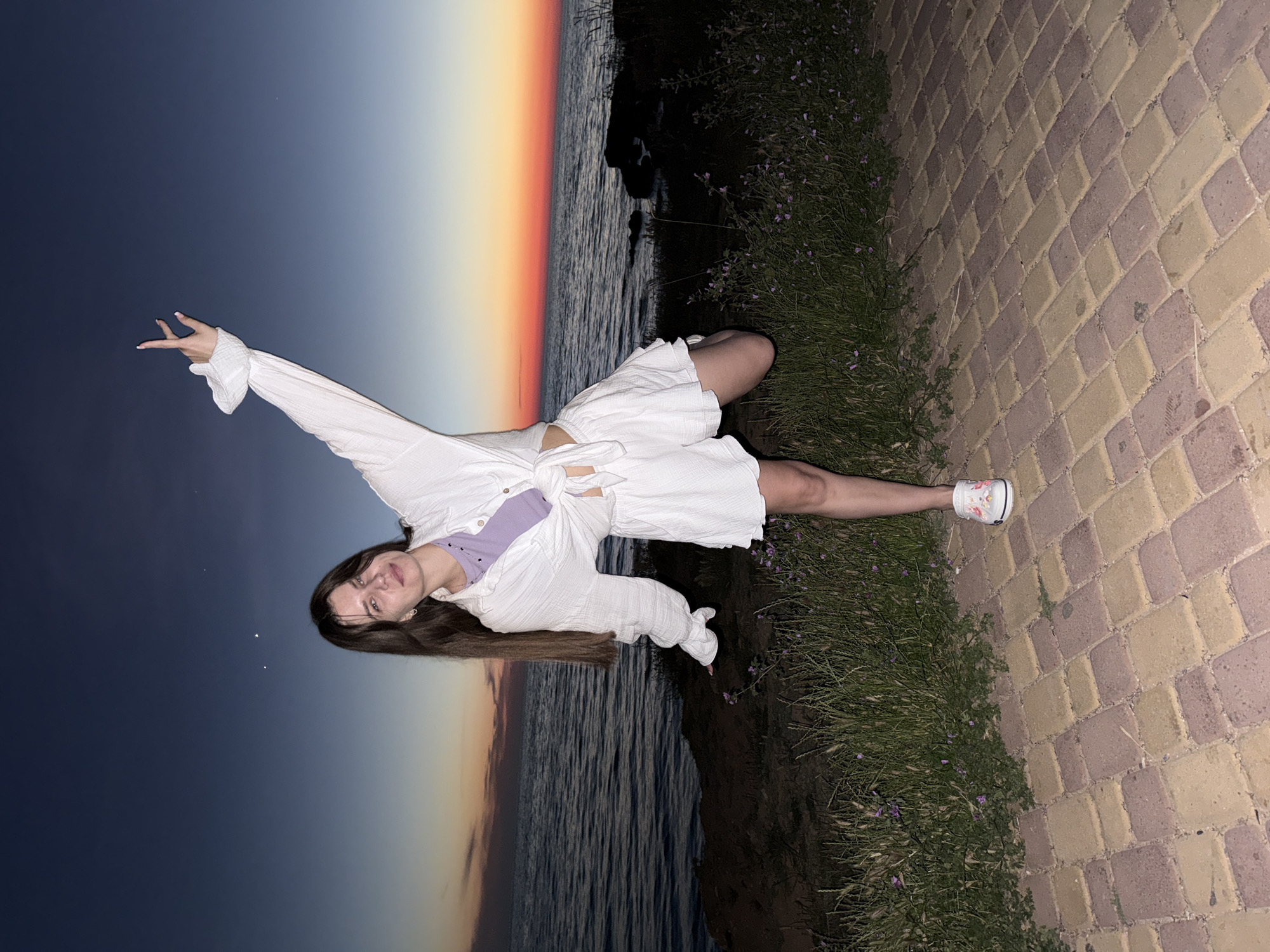 A young woman with long brown hair stands on a brick pathway near a body of water during twilight. She is wearing a white open shirt tied at the waist over a purple crop top, and matching white shorts. Her right leg is raised, and her right arm is extended upward with her hand making a peace sign. The sky shows a gradient of dark blue to orange, indicative of a sunset. The water is calm, and there is vegetation and rocky outcrops along the shoreline. The brick pathway is made of reddish-brown and yellow pavers.