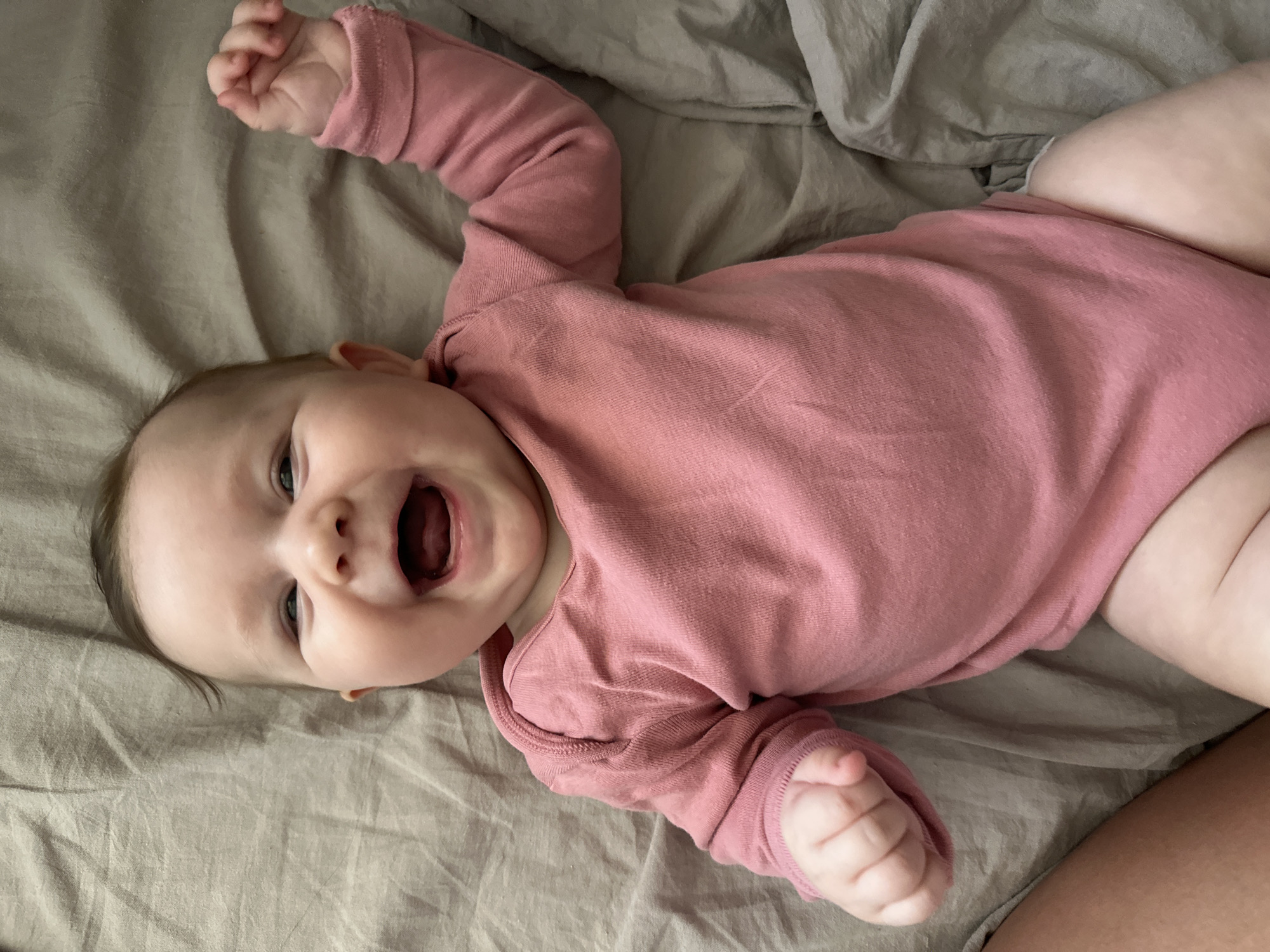 A baby wearing a pink bodysuit lies on a light brown sheet. The baby is on their back with their arms raised. Their mouth is open as if smiling or vocalizing. The location is not identifiable.