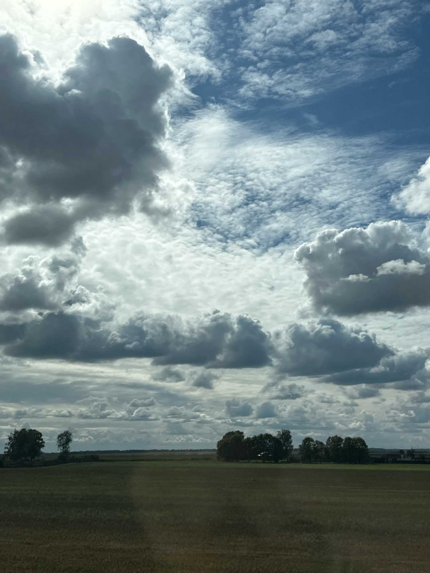 The image depicts a rural landscape under a dramatic sky. The foreground is dominated by a large, open field, likely agricultural, with a slightly textured surface suggesting stubble or dry grass. A line of trees separates this field from the horizon, with some scattered trees visible in the distance. The sky is a prominent feature, filled with a mixture of dark, heavy clouds and lighter, wispy formations, indicating changing weather conditions. The overall impression is one of vastness and natural beauty. The location is identified as Блонскі сельскі Савет, Belarus. There are no people or human activities visible in the frame.