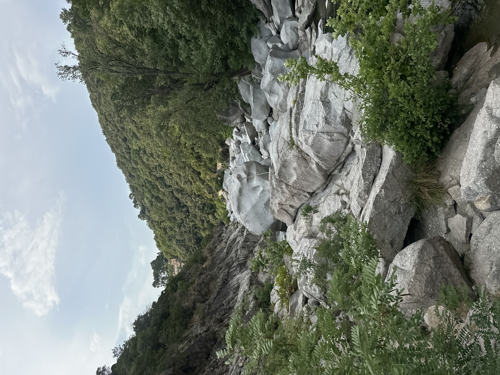 A wide shot captures a rocky riverbed with a stream flowing through it. The riverbed is filled with large, smooth, grey boulders of various sizes, some stacked atop each other, creating a rugged terrain. Lush green foliage and trees cover the steep hillsides that flank the river. The sky above is a soft blue with scattered white clouds. A building with a terracotta roof is visible in the distance, nestled among the trees on the hillside. The scene is natural and undisturbed, suggesting a remote or natural park location.