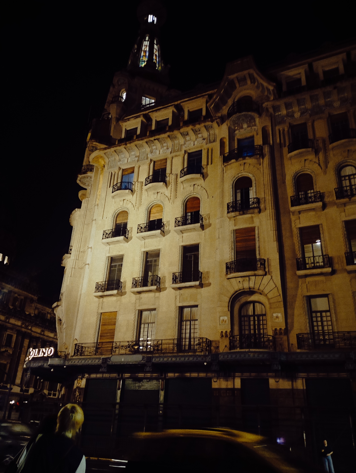 The image displays a nighttime urban scene centered on a multi-story architectural building at the intersection of Avenida Diagonal Norte (Roque Sáenz Peña) and Avenida Callao in Buenos Aires, Argentina, specifically the Palacio de la Prensa (ex-Hotel Majestic).
The dominant subject is a light-colored, ornate building, likely of Art Nouveau or eclectic design, illuminated by artificial light. Its facade features multiple levels of windows, many arched, equipped with dark metal decorative balconettes. Some windows show interior lighting, while others are dark or have closed wooden slat shutters. The building's summit is characterized by a distinct, illuminated dome or cupola featuring colorful stained-glass windows. The ground floor houses commercial establishments secured by dark roll-down shutters. To the lower left, a partial illuminated sign reads "OLINO".
The environment is dark, indicating night. The street in the foreground shows motion blur from vehicle lights, with a yellow blur suggesting a taxi. Two individuals are present: one person with light-colored hair is seen from behind in the lower-left foreground, facing away from the viewer. Another less distinct figure of a male stands on the far right, wearing a dark top and light pants, facing towards the left.