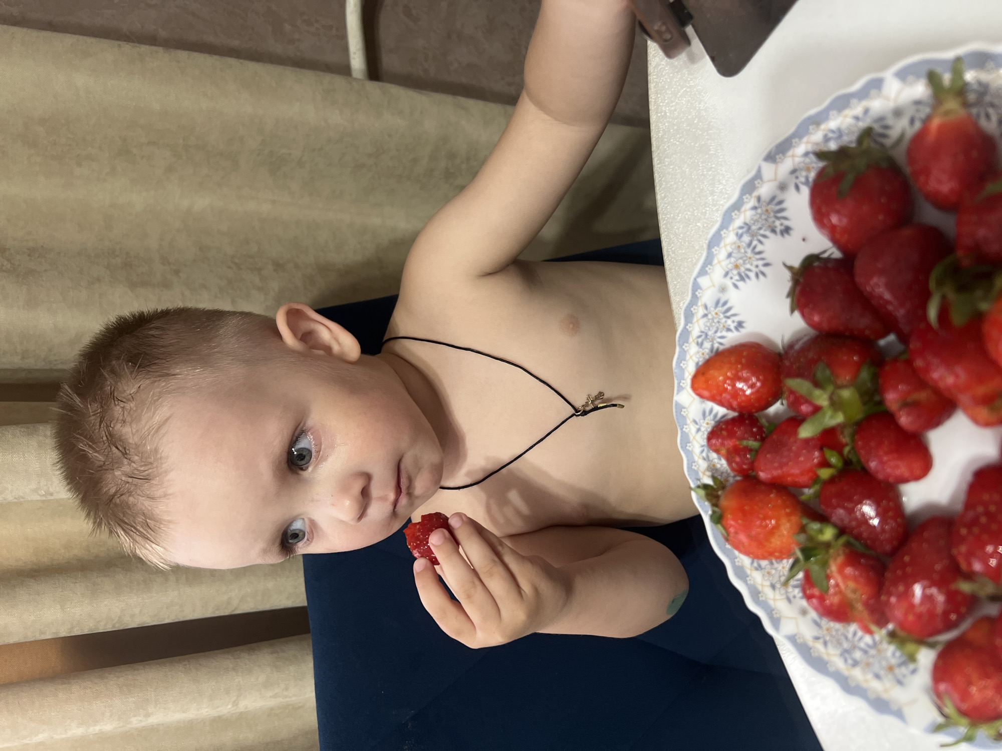 A partially clothed child, likely male, is seated at a table in what appears to be an indoor residential environment. The child is holding a strawberry in their left hand and has a plate of strawberries to the right of them on the table. They are wearing a necklace with a small pendant. A blue chair is visible beneath the child. Light brown blinds or curtains are in the background. While I do not have specific city context, the image comes from Марковский, Russia.