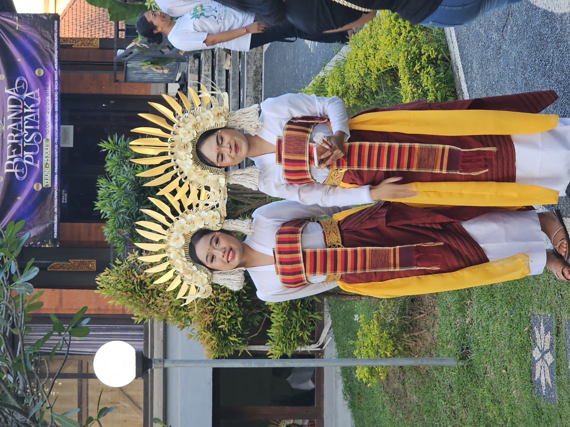 Two young women, dressed in traditional Balinese attire consisting of white tops, intricately patterned sashes, and maroon and yellow skirts, stand facing the camera in an outdoor setting. They wear elaborate golden headdresses with feathered embellishments and white tassels. The woman on the left smiles directly at the camera, while the woman on the right holds a smartphone in her hand and looks slightly to her left. Behind them, a building with a banner displaying "BERANDA PUSTAKA" and event dates is visible. The environment includes lush green foliage, a lamppost with a spherical light fixture, and a paved walkway. The scene suggests a cultural event or performance possibly taking place in Denpasar, Indonesia.