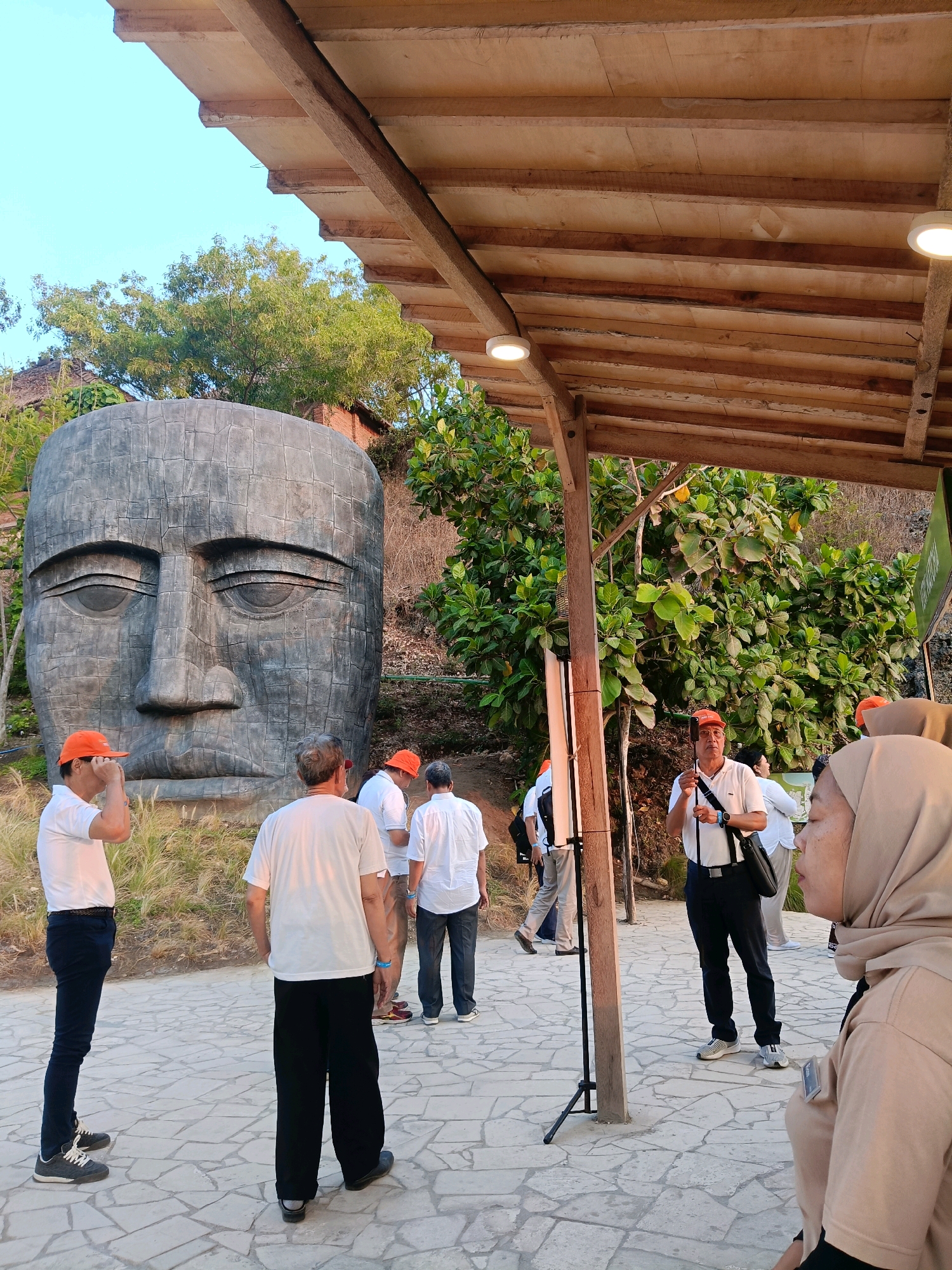 In Tanjungsari, Indonesia, a group of people are gathered outdoors near a large, stone sculpture of a human face. Several individuals, most wearing orange caps and white shirts, stand on a patterned stone pathway. One man in the foreground, wearing a beige hijab and a tan shirt, is facing right, partially obscuring another person. Behind the group, lush green trees and a hillside are visible. A wooden-framed structure with a wooden ceiling and lighting fixtures extends over a portion of the scene. The lighting suggests it is either early morning or late afternoon.