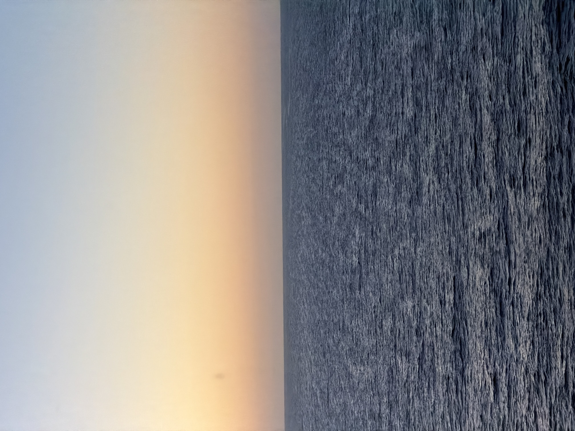 This media file presents a stark and minimalist landscape, divided vertically into two distinct halves. The left half of the image showcases a smooth, gradient sky, transitioning from a pale, cool blue-grey at the far left to a soft, warm orange-peach color towards the central dividing line. This subtle color shift suggests the natural light of either sunrise or sunset. The sky appears clear and cloudless, with no discernible atmospheric features.
The right half of the image displays a textured surface, predominantly in shades of deep blue-grey. This surface is characterized by numerous fine, mostly vertical lines and streaks, which evoke the appearance of rippled water, such as an ocean or a large lake, seen from above or at a distance. The texture implies subtle movement or currents on the water's surface.
A sharp, straight vertical line acts as the horizon, precisely separating the sky on the left from the textured water on the right. The image contains no visible people, objects, or specific events or activities taking place. The setting is an open, natural environment, likely a seascape. The notable details include the clear weather, the suggested time of day (sunrise or sunset), and the unusual vertical orientation of the horizon. The image conveys a sense of vastness and quiet contemplation. There is no visible text.