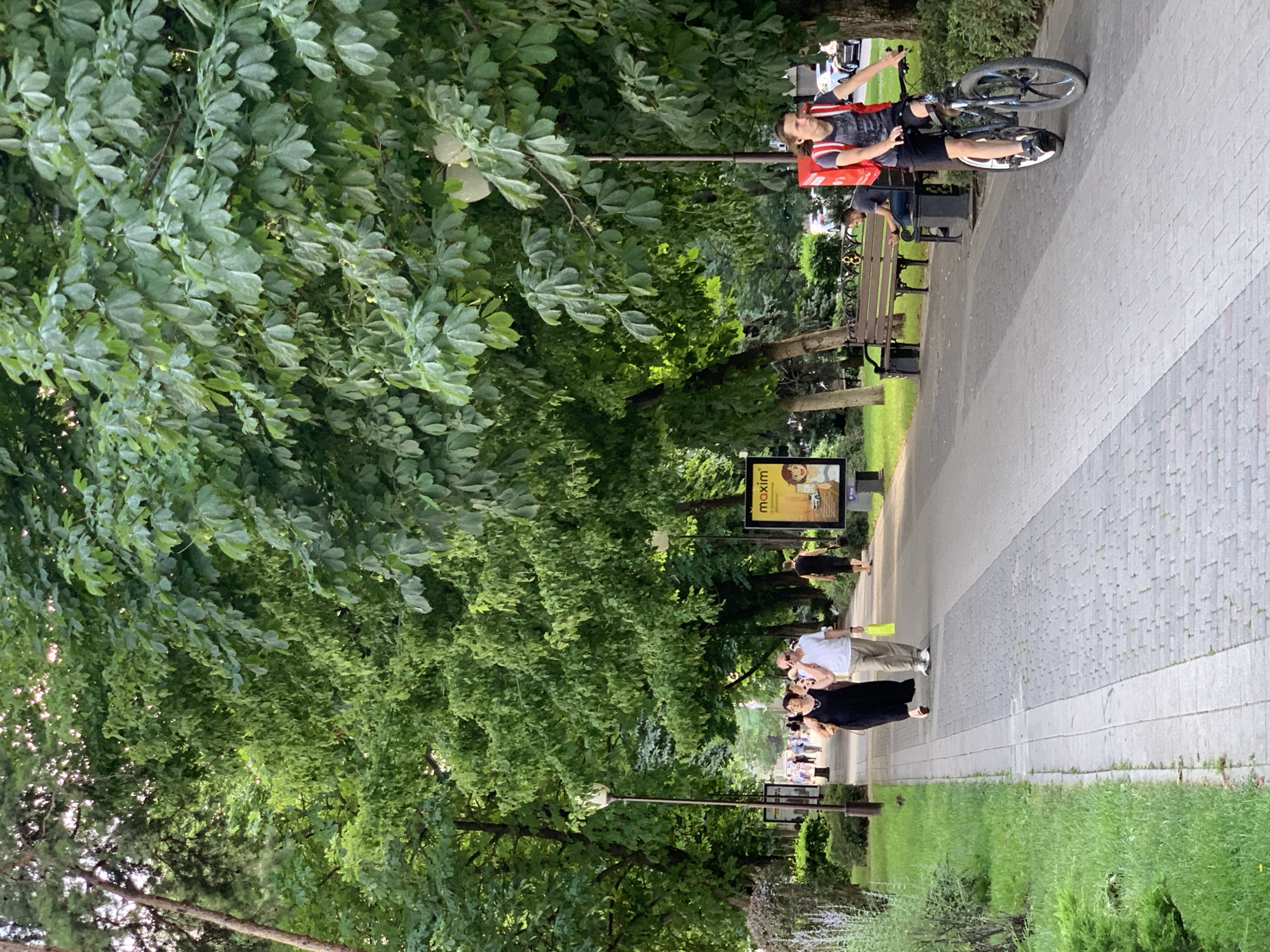 A person is riding a bicycle along a paved walkway lined with trees and benches. The person is wearing a black shirt with red accents and a red backpack, and they are looking to their left. Behind them, several other people are walking or sitting on benches. One person in the foreground, wearing a black dress, is looking towards the camera and smiling. Another person in a white shirt and beige pants is standing next to them.
A billboard with the text "maxim" and an illustration of two people is visible in the background. The time of day appears to be late afternoon or early evening, as the lighting is soft and the shadows are long. The weather seems pleasant, with green grass and full foliage on the trees. The scene is a public park or promenade.
