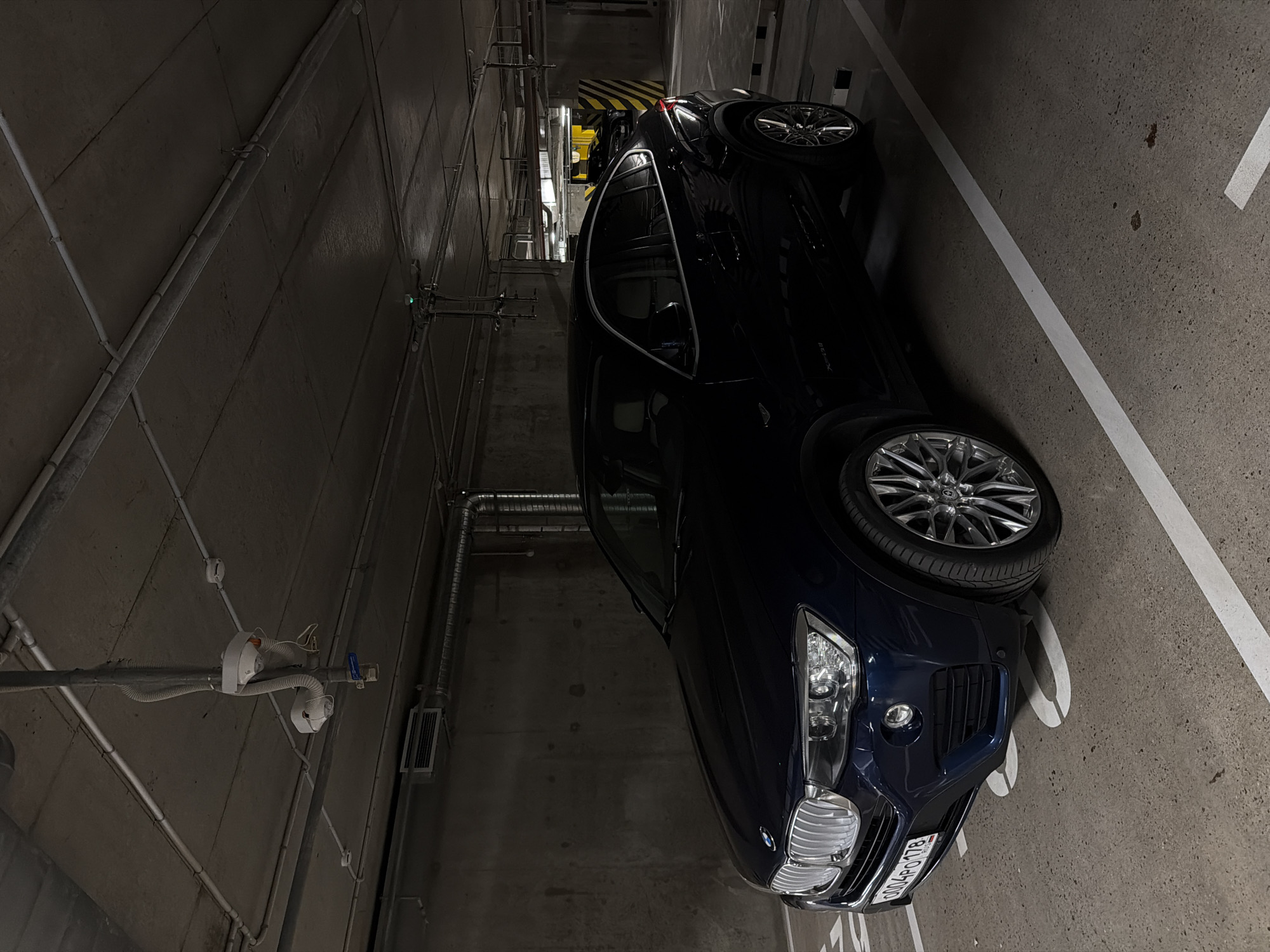 A dark blue BMW X6 is parked in an underground parking garage. The car is positioned in a parking spot marked with white lines. The garage has a concrete ceiling with exposed pipes and conduits. The walls are also concrete. The lighting is dim, typical of an indoor parking facility. The license plate of the BMW is visible and reads "0004PO 178 RUS". Another dark colored vehicle is visible in the background, parked diagonally in a yellow and black striped area.