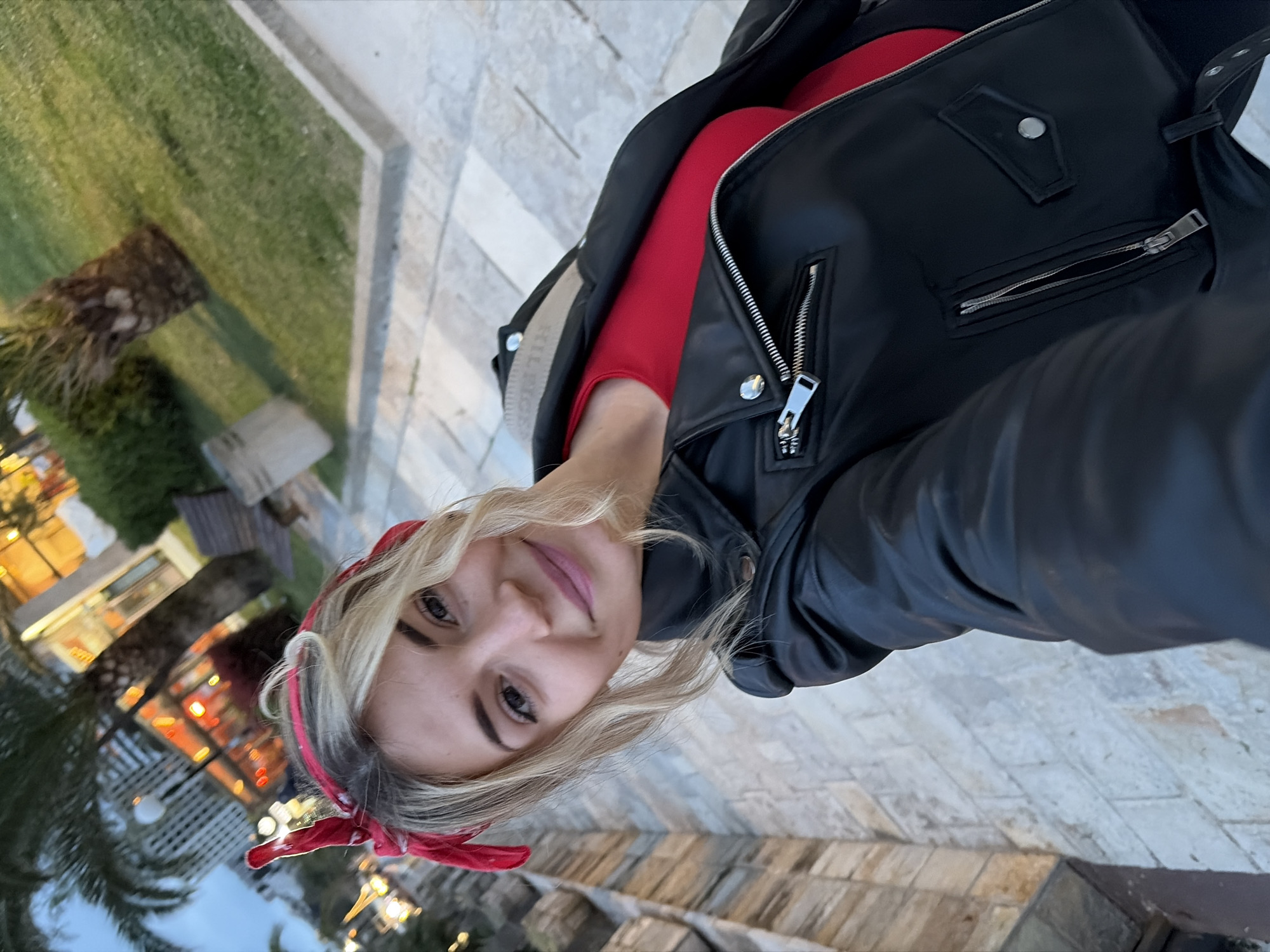 A selfie taken in Budva, Montenegro, shows a woman with blonde hair and a red bandana wearing a black leather jacket over a red shirt. The background includes a paved area, green grass, palm trees, and a building with illuminated windows. The photograph is slightly rotated.