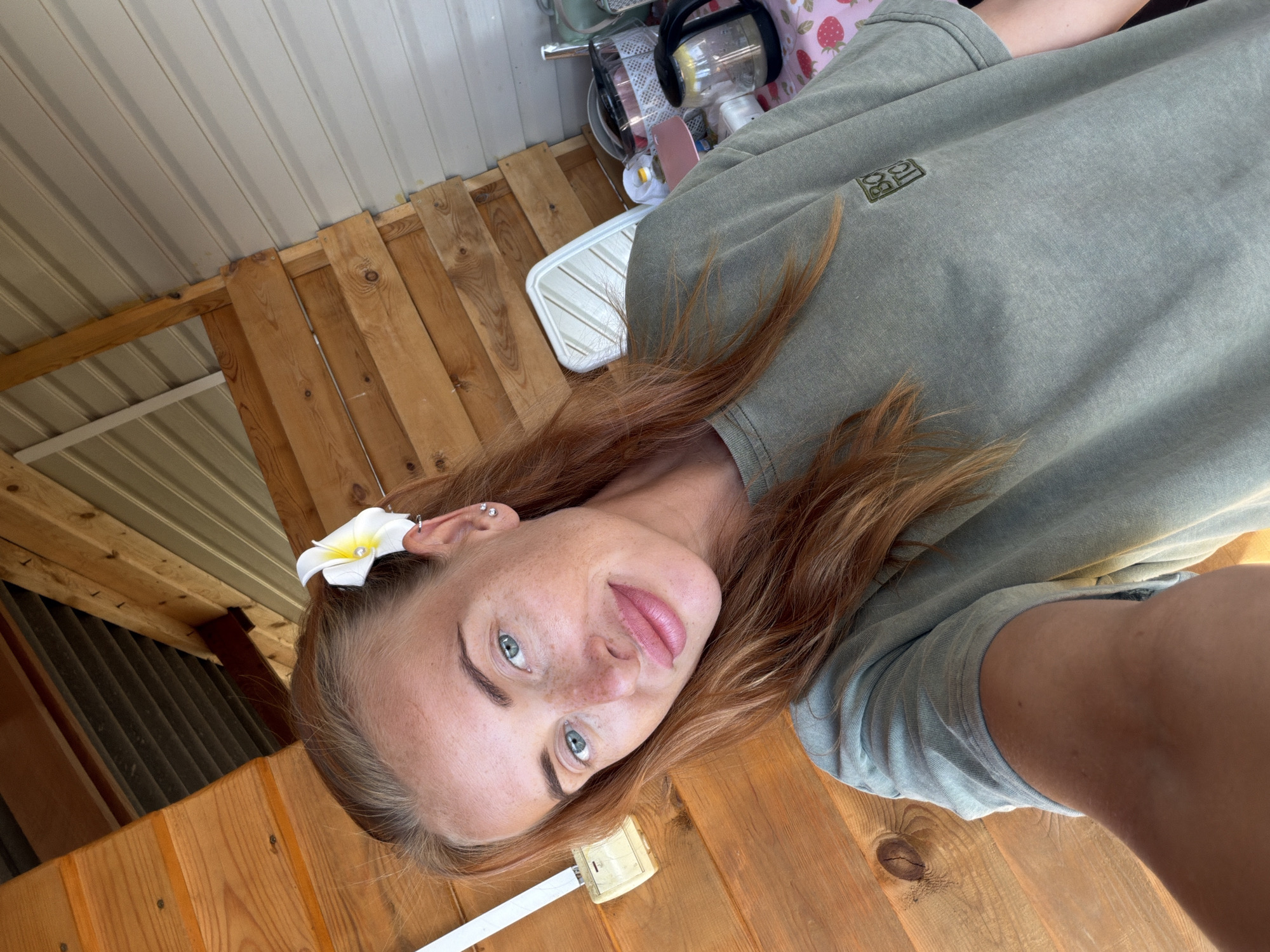 The image shows a young woman with red hair and blue eyes, lying on a wooden surface. She is wearing a green t-shirt with a small logo on the left breast. A white flower with a yellow center is tucked behind her ear. The background consists of wooden planks and a corrugated metal ceiling. There are some objects in the background, including a fan and what appears to be a blender, suggesting an indoor or covered outdoor setting, possibly a porch or shed. The woman is looking directly at the camera with a relaxed expression. The lighting suggests it is daytime.