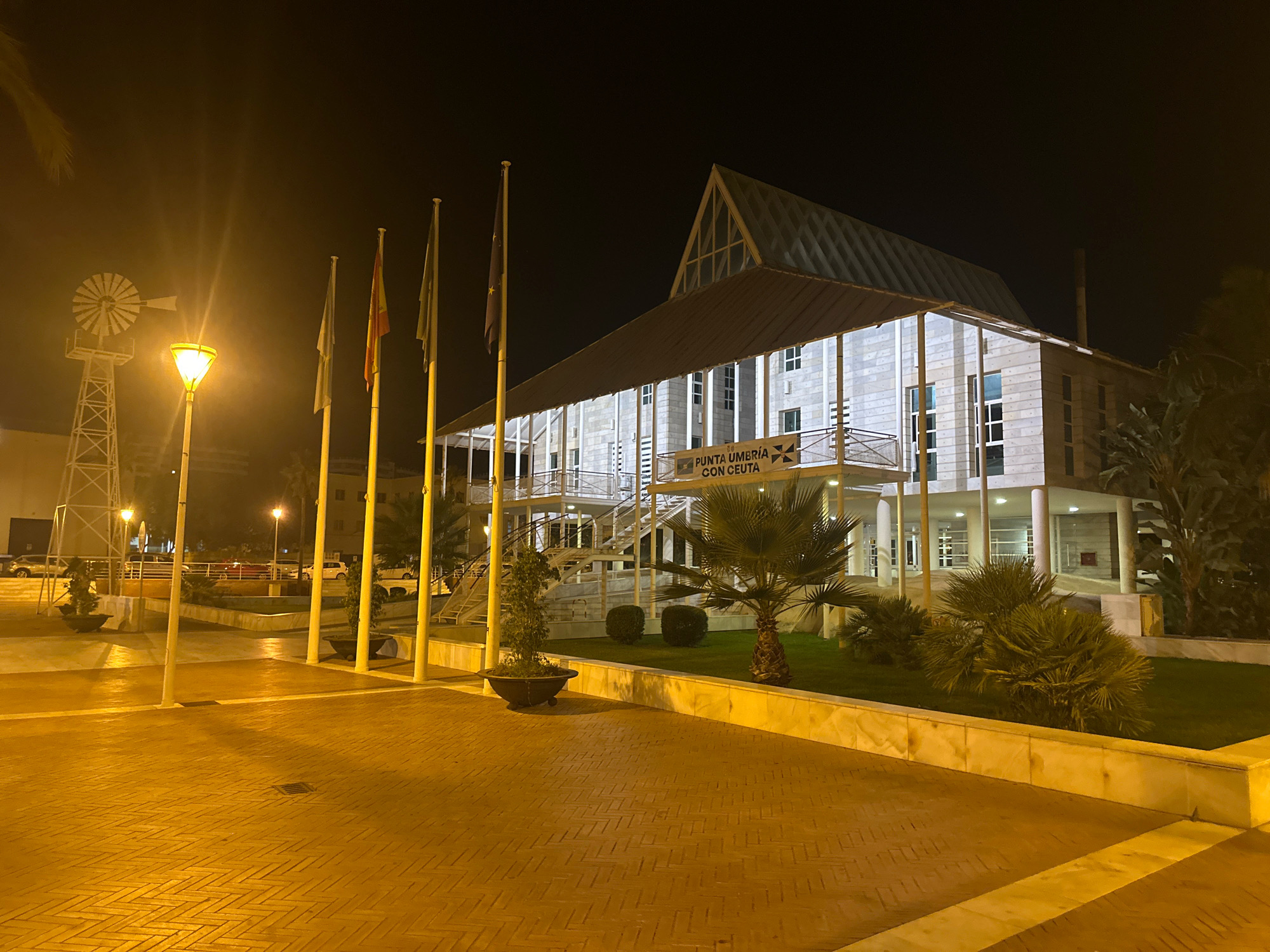 This image depicts a nighttime scene in Punta Umbría, Spain. The central focus is a modern, multi-story building with a distinctive peaked roof and extensive glass windows, illuminated by external lighting. The building's facade is constructed of light-colored stone or concrete blocks. A prominent sign above the entrance reads "PUNTA UMBRÍA CON CEUTA". In front of the building, a wide, brick-paved walkway is illuminated by a street lamp. Several flagpoles with flags are positioned on the walkway. To the left, a tall, lattice-like structure with what appears to be a windmill at the top is visible, also illuminated. The surrounding area includes manicured greenery, palm trees, and bushes, with a low marble or stone wall bordering a grassy area. In the background, parked cars are visible. The overall atmosphere is one of a well-lit public space at night.