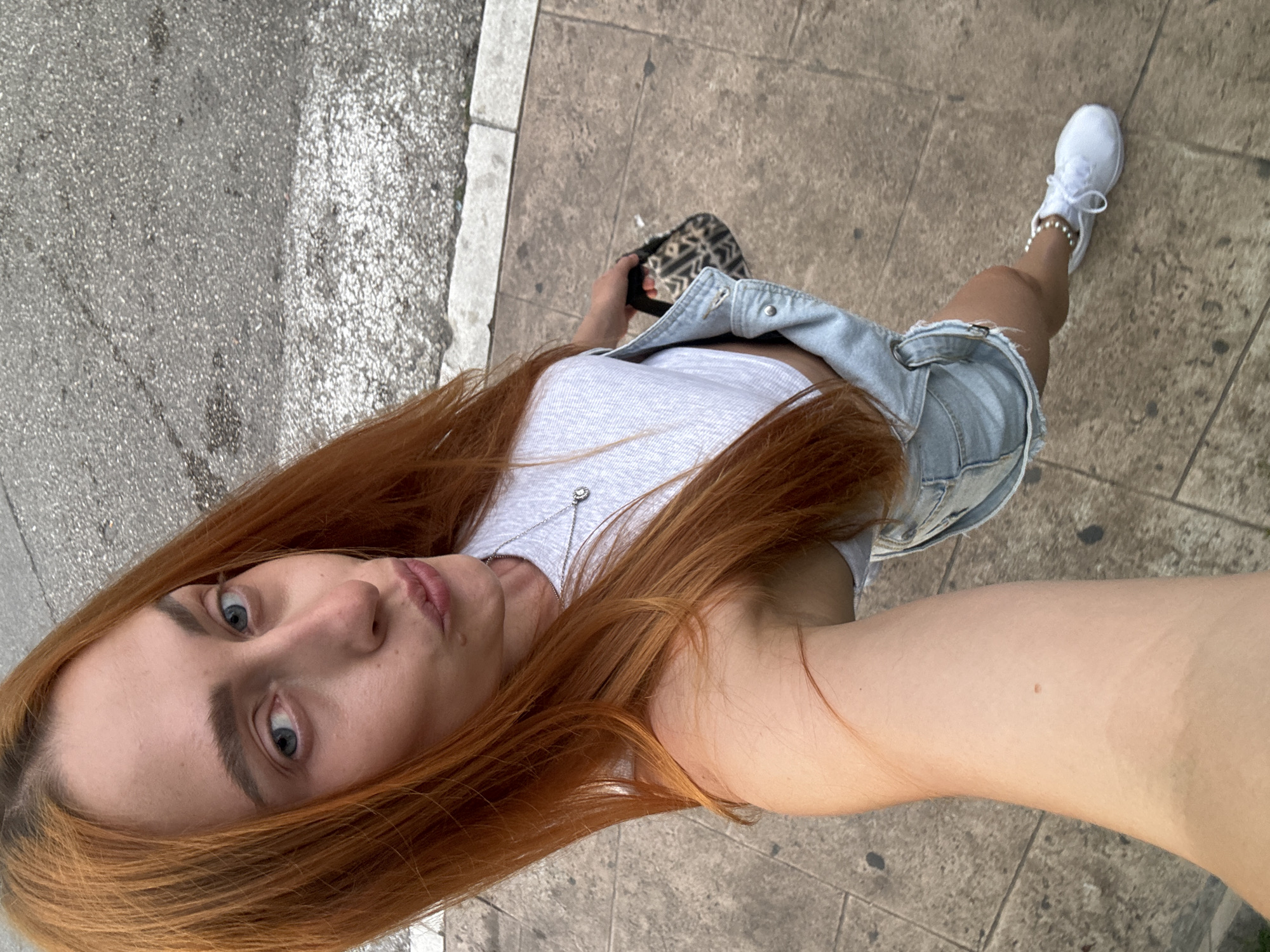 A high-angle, full-body selfie taken from a slightly upward perspective shows a young, fair-skinned woman with long, auburn hair and blue eyes. She is wearing a white crop top, denim overalls, and white sneakers with white laces. Her left arm is extended to take the photo, and she is holding a small, patterned clutch bag in her right hand. She is standing on a paved sidewalk, with a street visible in the background. The image appears to have been taken outdoors, likely in a city setting, possibly Budva, Montenegro, given the architectural style of the paving stones and the general appearance of the street.