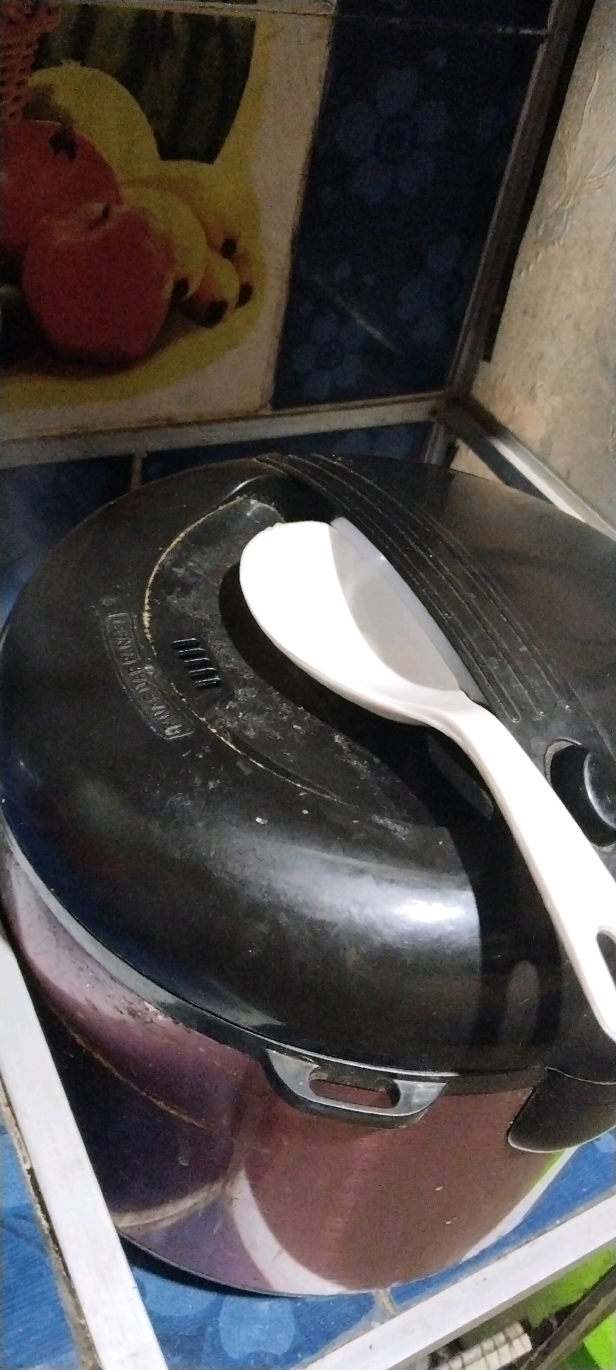 A white plastic rice paddle rests on the black lid of a rice cooker. The rice cooker has a maroon base with a metallic sheen and is situated on a surface with a blue tiled pattern. In the background, there is a wall decorated with a colorful image of fruits, including what appears to be an apple and bananas, against a dark, patterned wallpaper. The scene is set indoors, likely in a kitchen, in Tanjung Ratu Ilir, Indonesia.