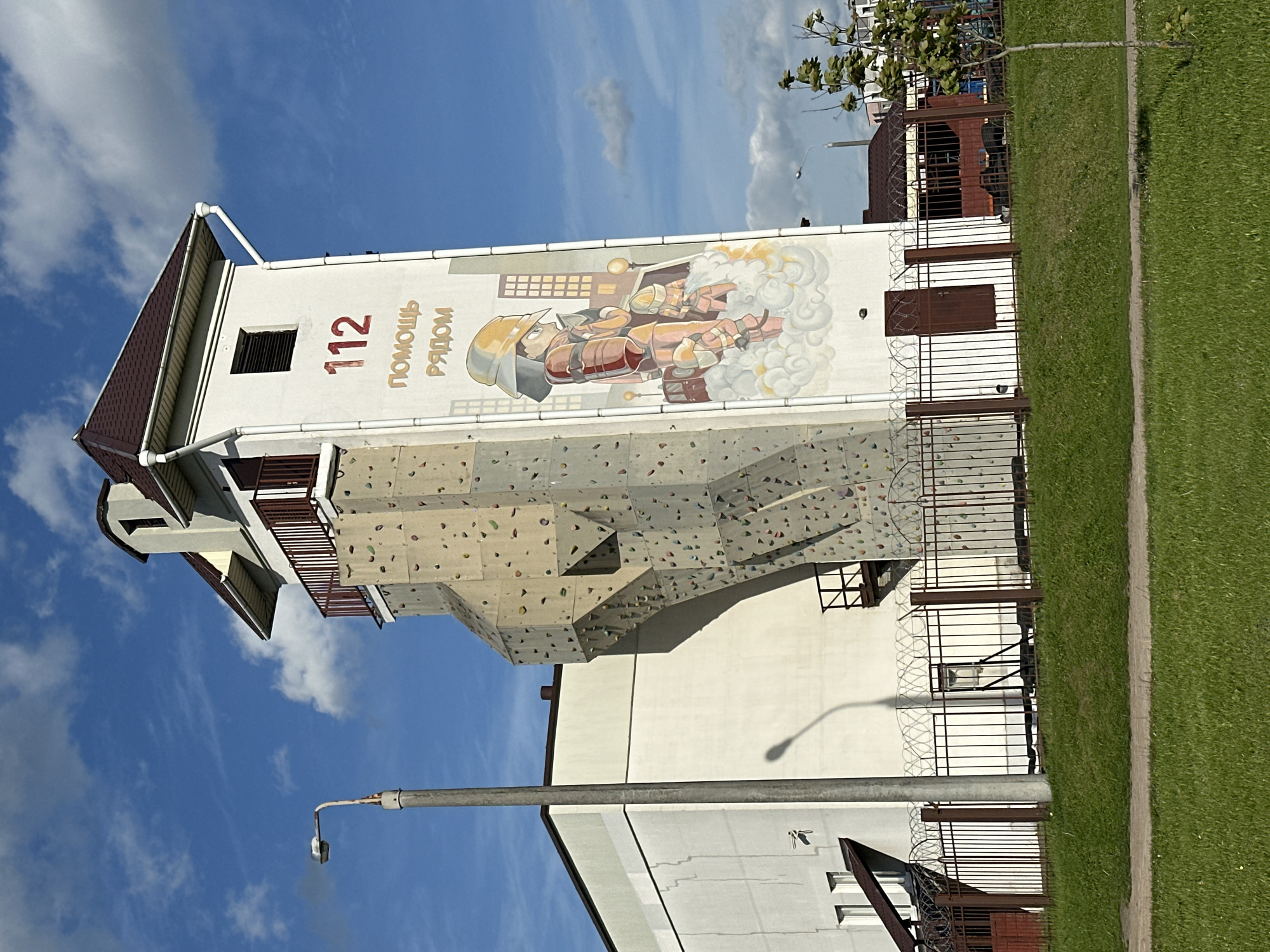 The image shows a building in Minsk, Belarus, that appears to be a fire station or emergency services facility. The building has a tall tower structure featuring a climbing wall on one side and a mural depicting a firefighter on another. The mural includes the number "112" and the phrase "ПОМОЩЬ РЯДОМ", which translates to "Help is near" in Russian. The building is surrounded by a fence and some vegetation, including grass and trees. The sky is blue with some scattered clouds. A light pole stands nearby, and other buildings are visible in the background.