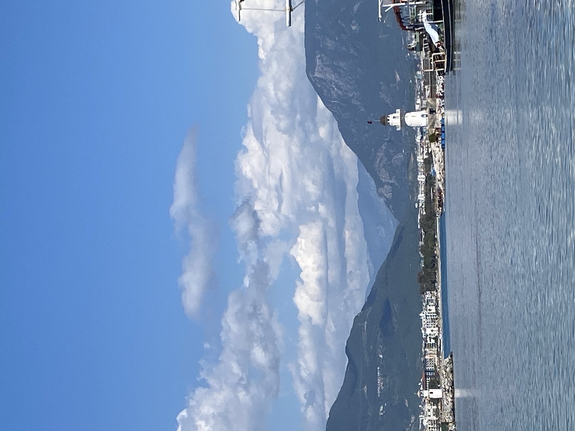 The image captures a scenic view of Alanya, Turkey, featuring a clear blue sky with scattered white clouds. Mountains rise in the background, their slopes covered with vegetation. Along the coastline, there's a stretch of buildings, predominantly white, indicating a coastal city or resort area. The sea is calm, reflecting the sky. A lighthouse is visible along the coast. A docked boat can be seen in the harbor.