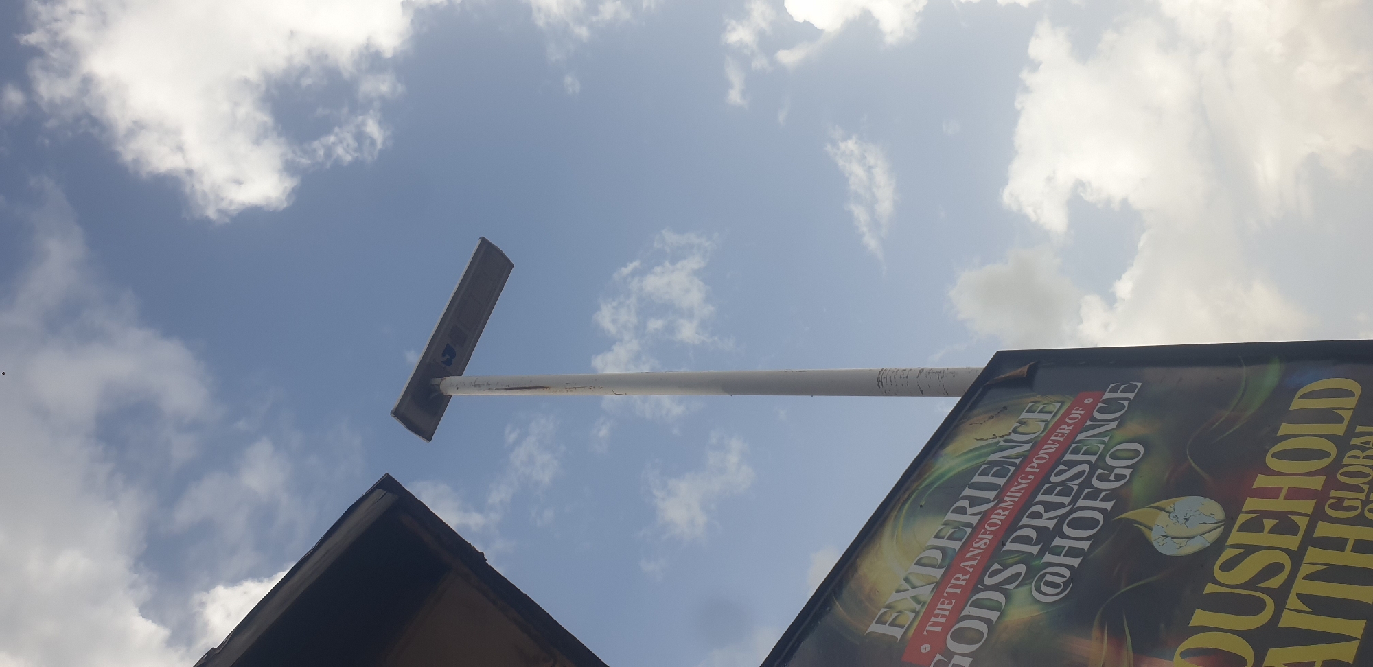 The media file shows a low-angle view of a street light pole against a bright, cloudy sky. The pole is white and extends horizontally from the left of the frame towards the right, with a dark, rectangular fixture at its end. Below and to the right of the pole, a portion of a large billboard is visible. The billboard is dark with vibrant graphics and text.
The visible text on the billboard reads: "EXPERIENCE THE TRANSFORMING POWER OF GOD'S PRESENCE @HOFGOCULT" and "HOUSEHOLD FAITH GLOBAL". The billboard appears to be an advertisement for a religious event or organization.
The sky is blue with scattered white, fluffy clouds, suggesting it is daytime, likely midday or afternoon due to the brightness. The overall scene appears to be outdoors, possibly on a street or in an open area in Barakin Lerawi, Nigeria, as indicated by the location context. There are no people visible in the image. The angle and the presence of the billboard suggest an urban or semi-urban setting.